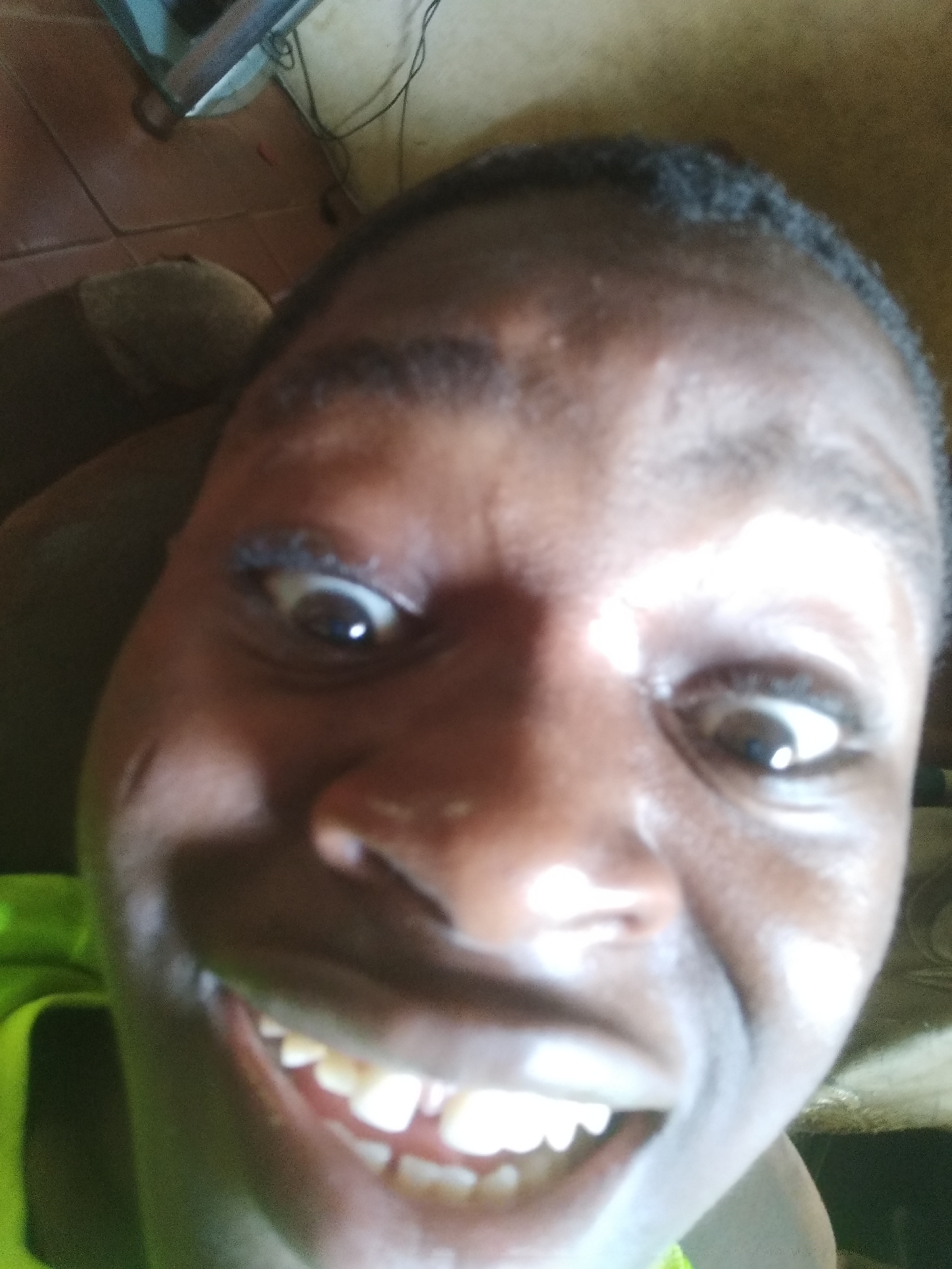 A close-up selfie of a Black male with dark skin and short, dark hair. His eyes are wide and directed upwards. He is smiling, exposing his teeth. The background includes parts of a room with tiled flooring. The location is Shangisha, Nigeria.