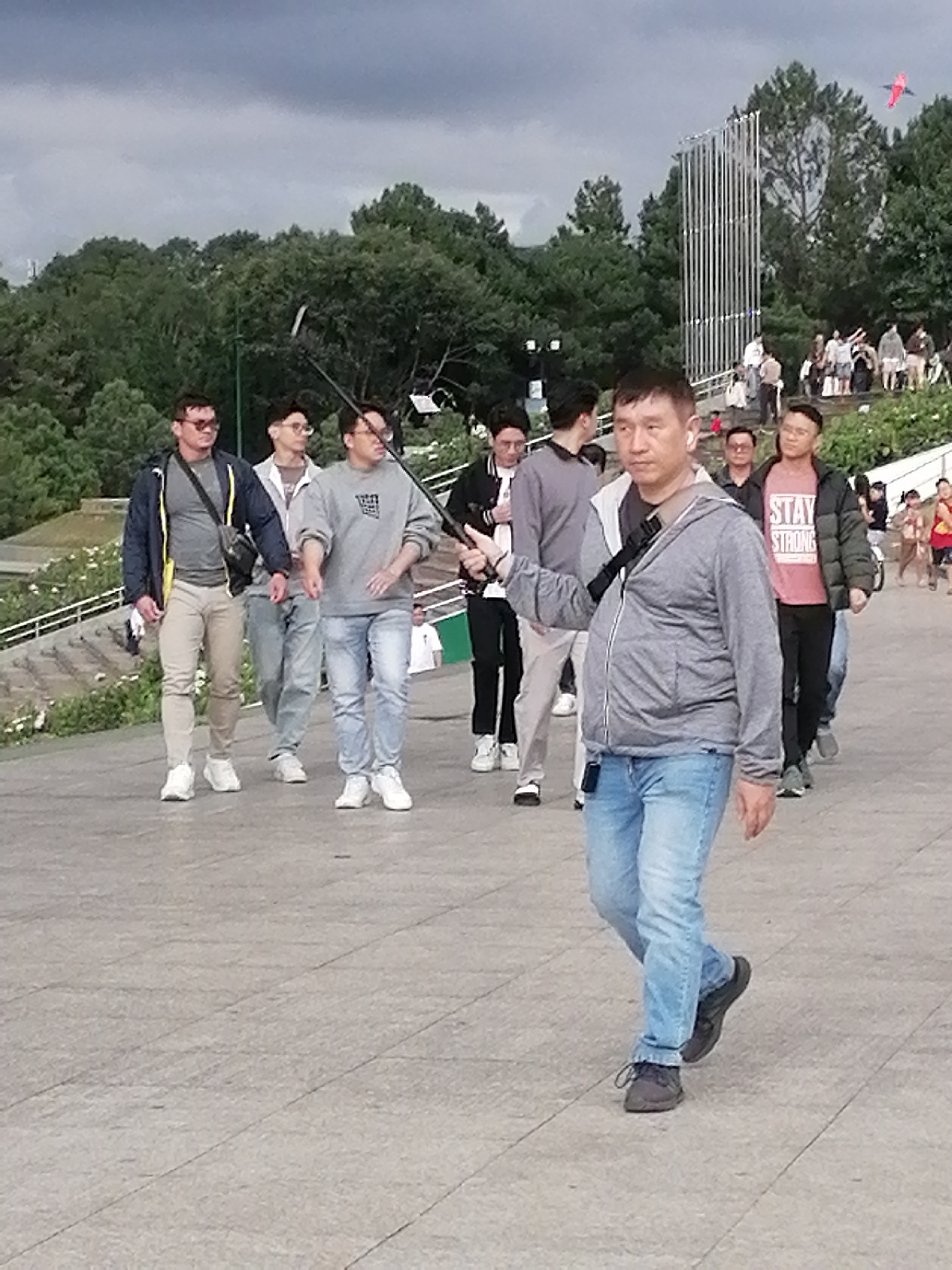 A group of people are walking on a paved path in a park. The path is grey and textured, with visible grout lines. In the background, there are lush green trees and a large, modern, white metal sculpture with vertical slats. The sky is overcast and grey, with a hint of a red kite visible in the upper right corner. The people are casually dressed in t-shirts, hoodies, and jeans. Some are walking together, while others are more solitary. One man in the foreground is holding a selfie stick. The scene appears to be outdoors in a public space, likely a tourist attraction or recreational area. The presence of the sculpture and the overall landscape suggest a city park. This park is Xuan Huong Lake in Da Lat, Vietnam, famous for its recreational activities and scenic beauty.