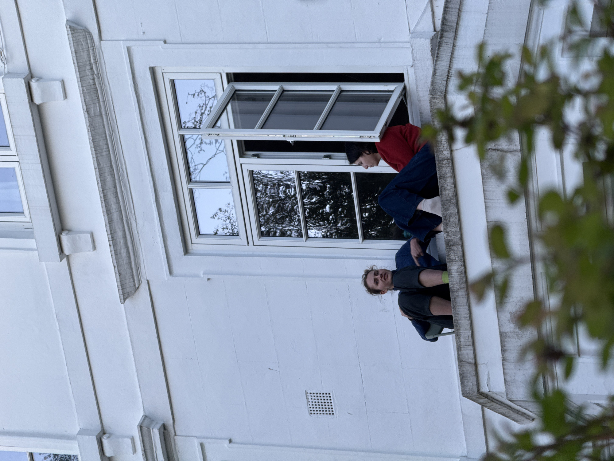 Two people are visible on the exterior of a white building with a large window. One person, wearing a red shirt and blue jeans, is leaning out of the window. The other person is sitting on the window ledge, wearing a gray shirt. The building appears to be located in London, United Kingdom.