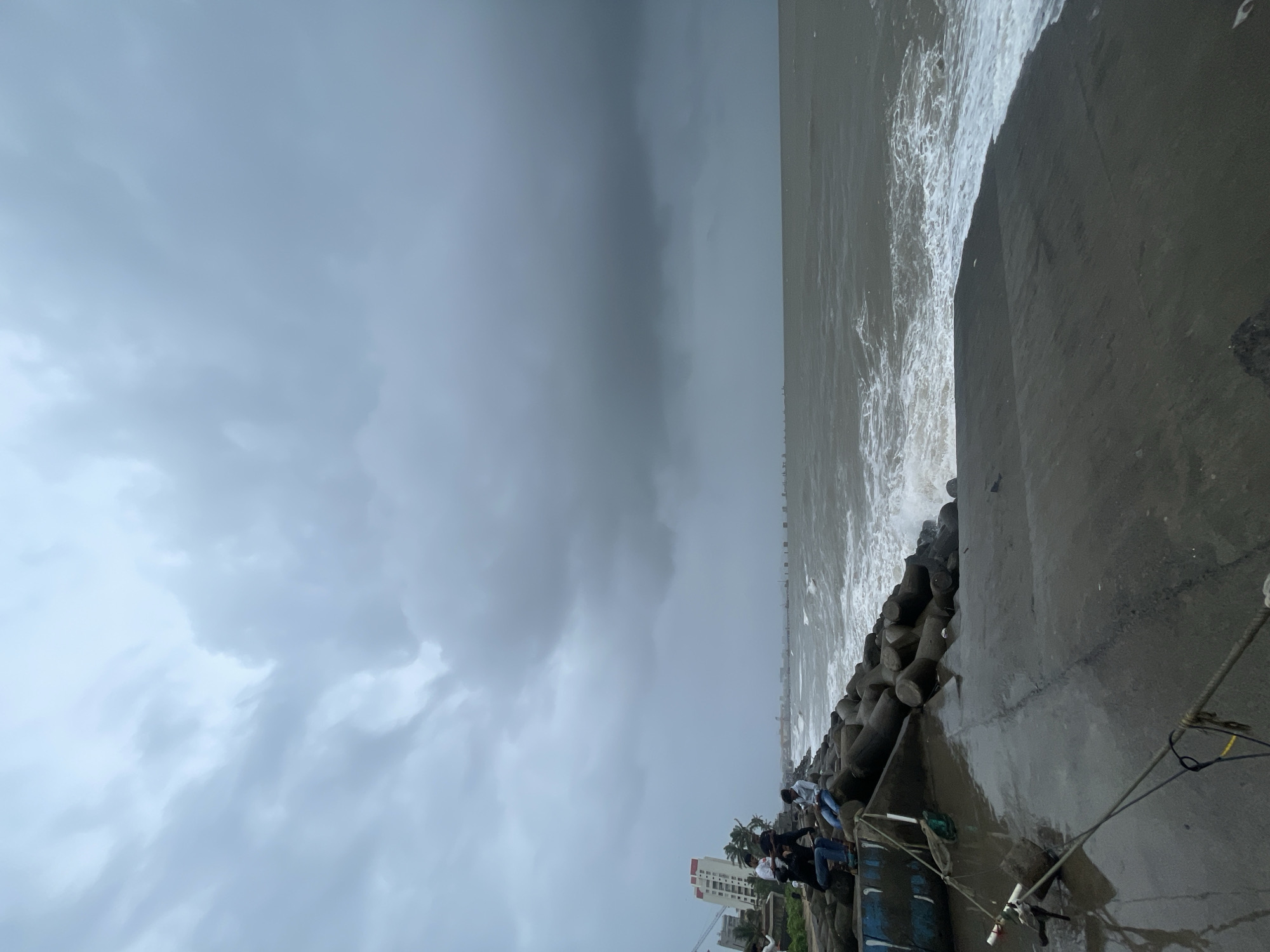 The image depicts a cloudy, overcast day at Bandra Fort, also known as Castella de Aguada, in Mumbai, India. The foreground shows a concrete walkway that is wet from recent rain or sea spray, with a rope tied to what appears to be a fishing rod assembly. To the left, a blue and white boat is partially visible, also wet. A line of large, concrete tetrapods form a breakwater along the coastline, with waves crashing against them. Several young men are seated and standing on the tetrapods, observing the turbulent sea. In the background, a tall white building with red accents and a construction crane are visible, indicating an urban setting. The sea is rough, with whitecaps and foam indicating strong winds. The sky is filled with heavy, dark gray clouds, suggesting impending rain or a storm. The overall atmosphere is moody and dramatic, typical of the monsoon season in Mumbai.
