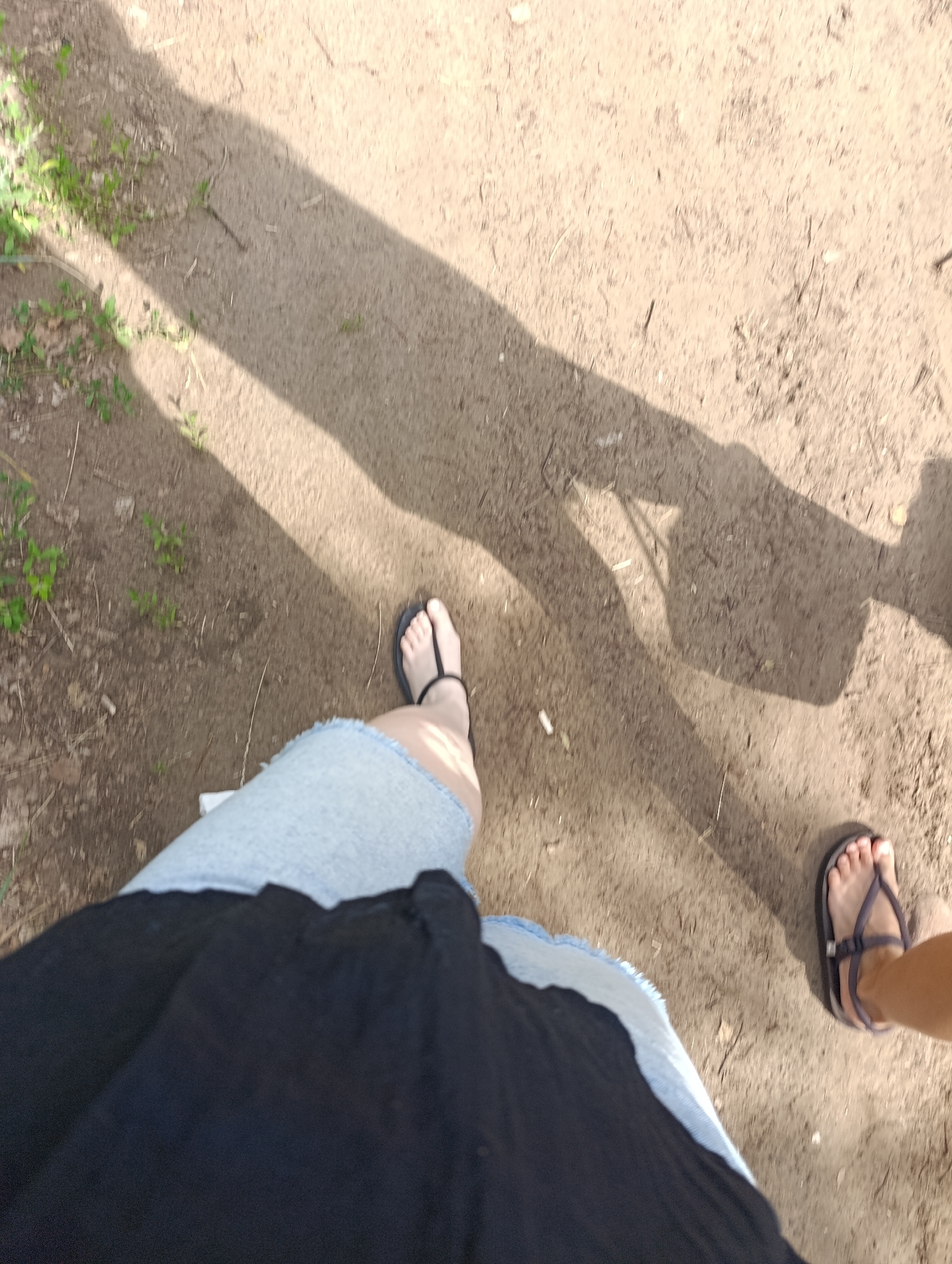 The image shows a first-person perspective of someone walking on a dirt path. The path is dry and dusty, with a few sparse blades of grass and some scattered twigs. A long shadow is cast across the path, suggesting it is daytime, likely late afternoon or early morning given the angle of the shadow. The shadow indicates the presence of at least one other person walking ahead.
The subjects are two individuals' legs and feet, both wearing casual footwear. One person is wearing dark flip-flops and has their left foot and lower leg visible. They are also wearing a light blue denim skirt with a frayed hem and a dark black fabric, possibly a dress or top, that extends down from the waist. The other person's right foot and lower leg are visible further ahead and to the right, wearing dark-colored sandals with thin straps. Their leg is tanned and appears to be wearing shorts or a skirt.
The scene appears to be outdoors in a natural, unpaved area. The lighting is bright, indicating sunny weather. There are no discernible emotional cues, as the focus is on the lower bodies and the ground. No text is visible in the image.