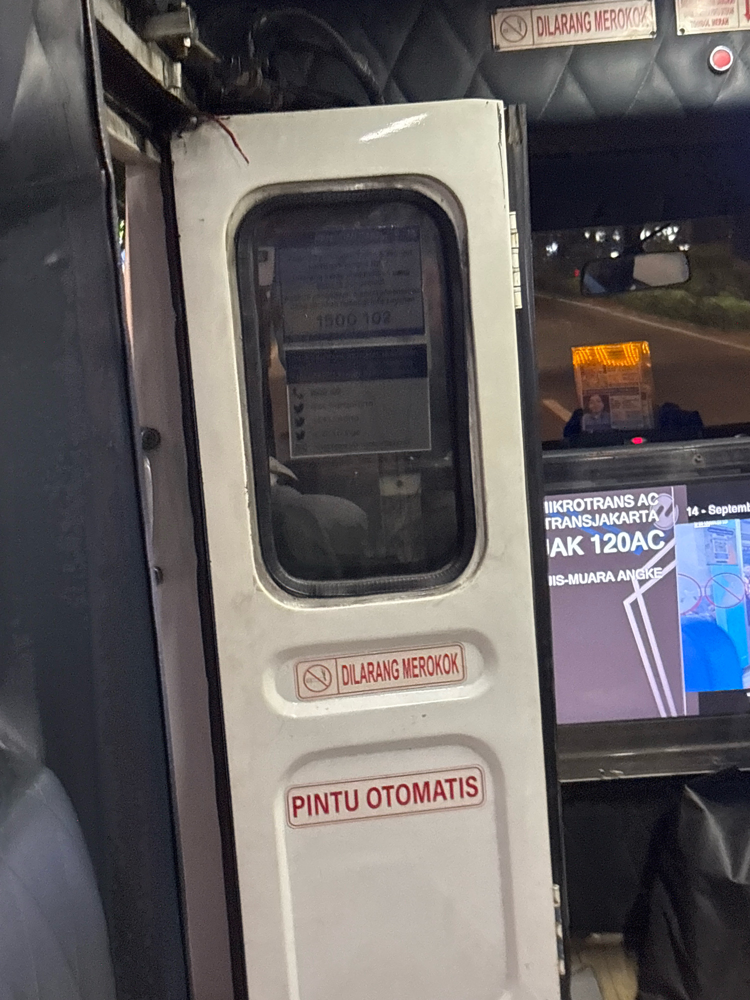 An interior view of a bus door in North Jakarta, Indonesia, shows a white door with a window. A sign with a red border and white text reads "DILARANG MEROKOK" (No Smoking), and below it another sign reads "PINTU OTOMATIS" (Automatic Door). The window shows a reflection of the street outside and an advertisement on a screen. The bus appears to be stationary.