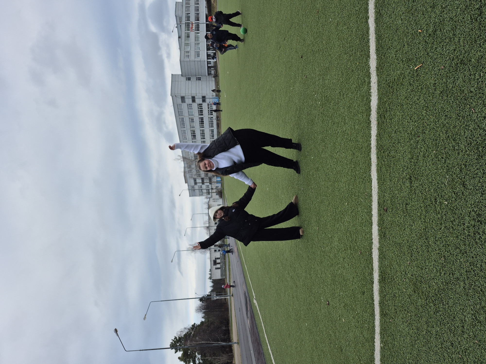 This outdoor photograph, rotated 90 degrees counter-clockwise, depicts an artificial turf sports field and its surroundings under an overcast sky in Chaikovsky, Russia.
In the foreground, two young women are playfully posing on the green artificial grass, holding hands. The woman on the left, wearing a white long-sleeved top, black pants, and a black vest, smiles at the camera with her arms extended outwards. She supports the second woman, who is dressed in a black jacket, black pants, and a light-colored hat or hoodie, leaning backwards in a dramatic pose with her arms also outstretched and a slight smile on her face. Her shoes appear to be brown.
Behind them, further down the field, several other individuals are visible. A group of children or young people are engaged in playing soccer with a bright green ball; one child wears an orange/red hat. Other figures are scattered across the field, suggesting ongoing activity.
Beyond the field, a paved path or road runs parallel to the turf, with streetlights visible along its edge. In the background, several multi-story buildings, likely residential, line the horizon. Trees, some appearing bare, are also present. The sky is uniformly gray and cloudy, indicating an overcast day, and the clothing suggests cool temperatures. No visible text is present in the image.