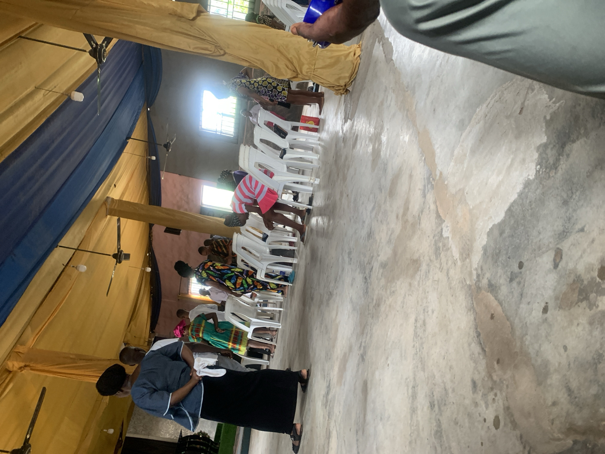 A photograph captures a scene inside a church located in Aba, Nigeria. A row of women in casual attire bow their heads, resting them on the backs of white plastic chairs. The walls of the building appear to be painted in pink hues with visible windows. The ceiling is lined with yellow and blue fabric; several ceiling fans are mounted. The floor of the room is concrete. People are milling around the room in the background.