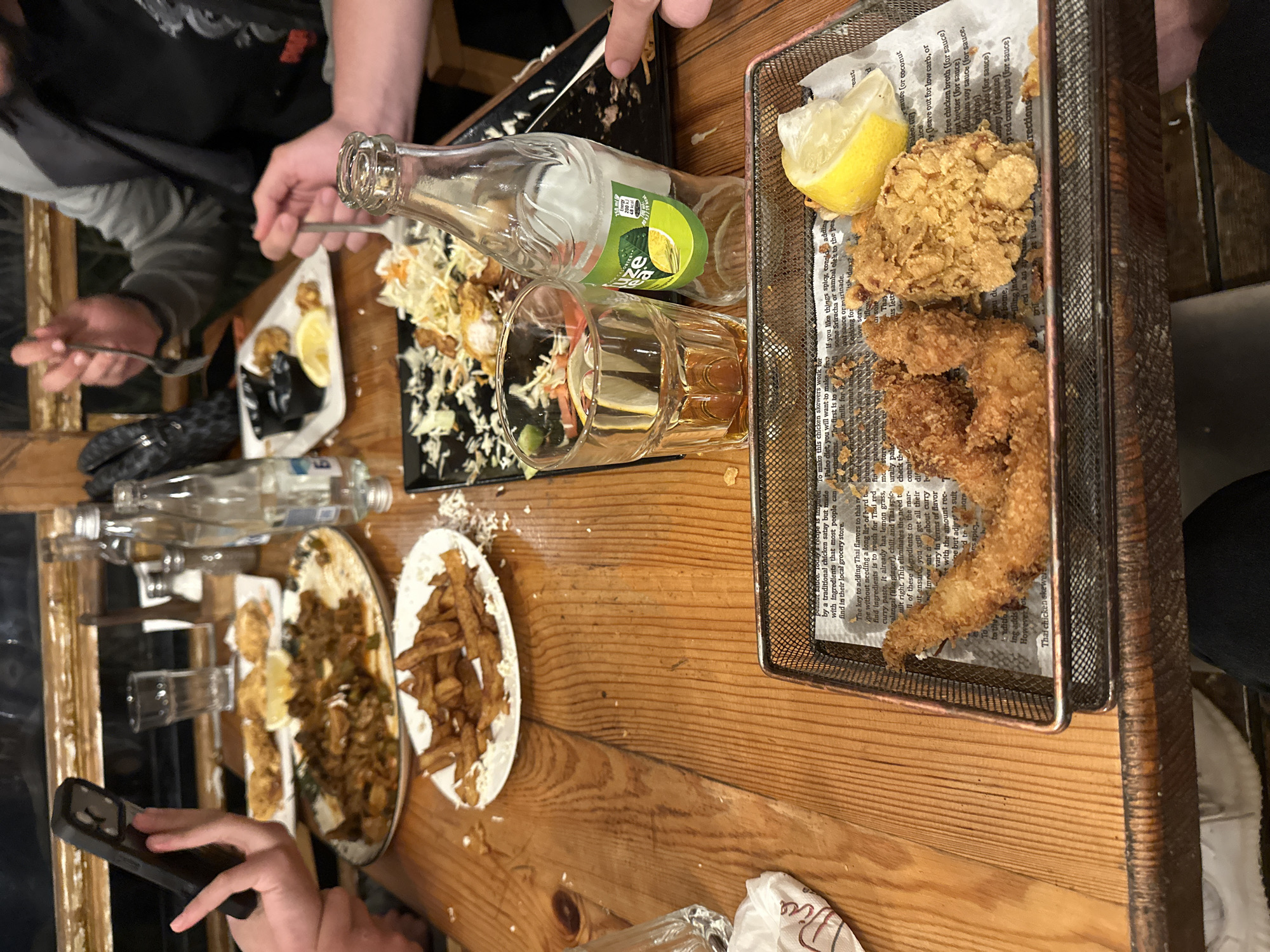 The image depicts a table at a restaurant, with various food items scattered across its surface. Visible are plates of fried chicken, fries, and a salad with dressing. Drinks such as a bottle of Sprite and glasses of amber liquid are also present. People are seated around the table, some holding utensils and phones, suggesting they are either eating or taking photos of the food. The location in the city of Blagoevgrad, Bulgaria, cannot be determined based on the provided information.