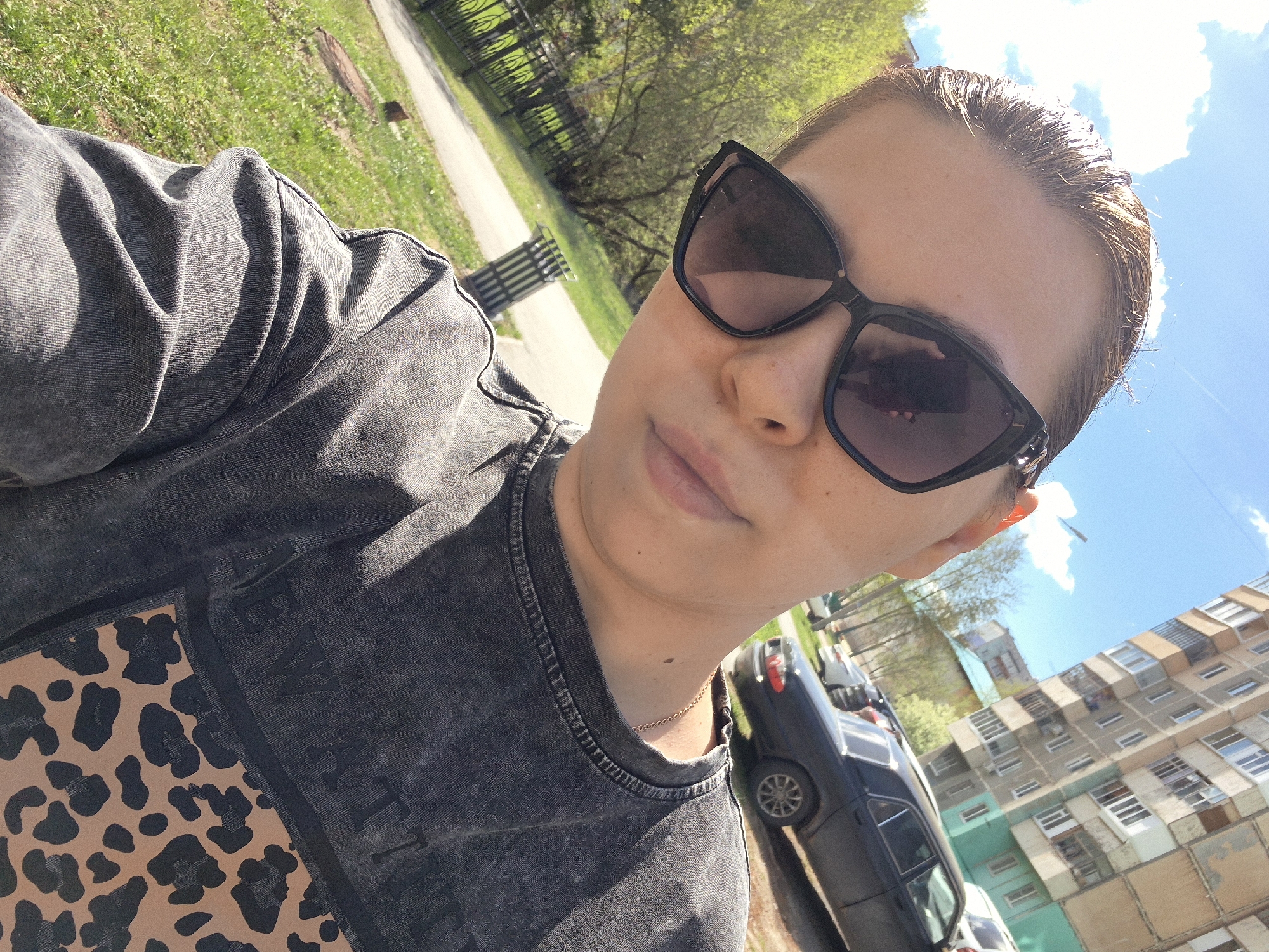 A woman is taking a selfie outdoors. She wears sunglasses and a gray shirt with a leopard print design. A gold chain is visible around her neck. Behind her are trees, a sidewalk, a black fence, and a park bench. A black car is parked nearby, and a tall residential building with rows of windows is in the background. The location appears to be in a city, possibly Tobolsk, Russia. The sky is blue with some clouds.