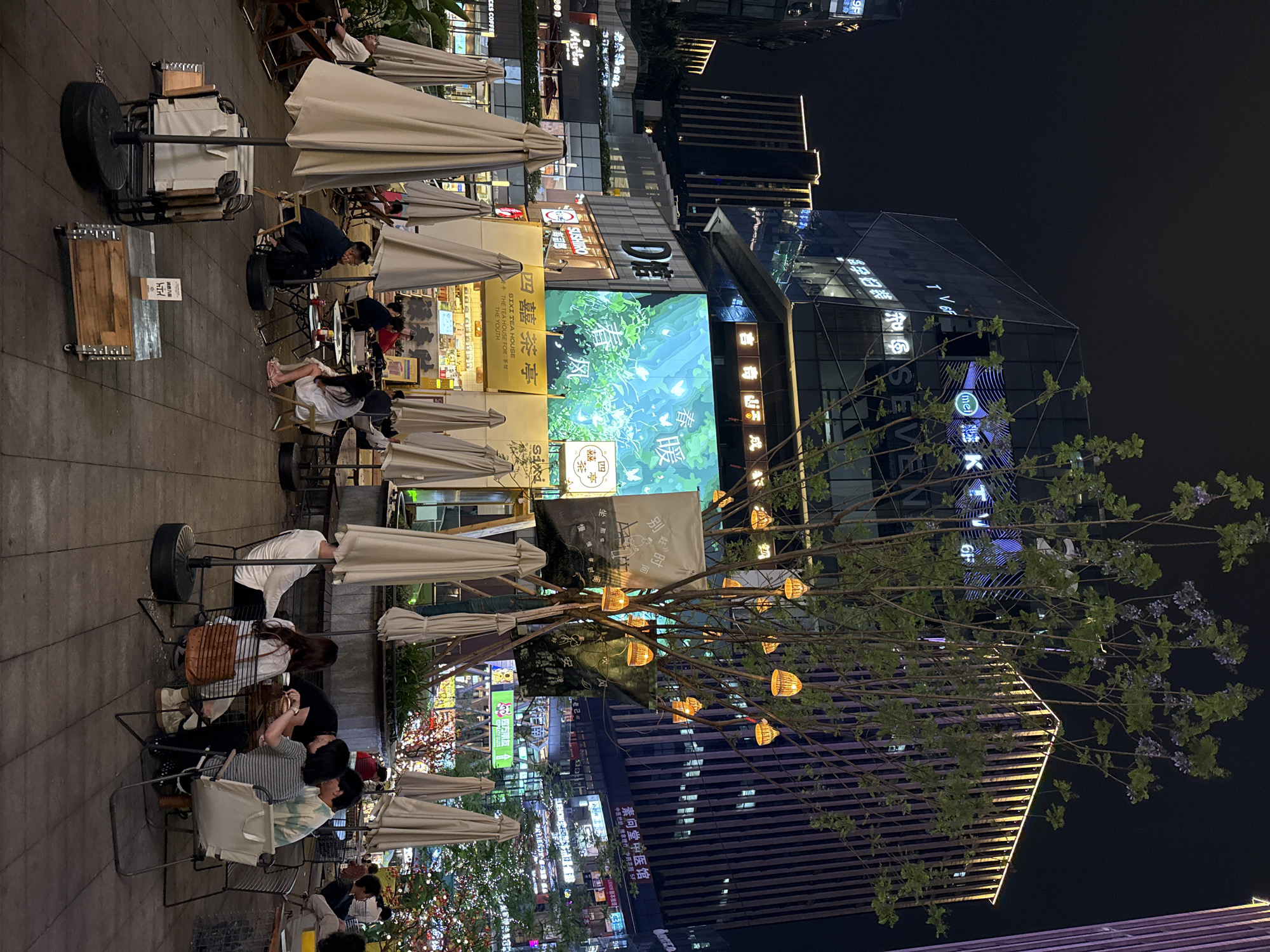 A nighttime scene in the Yuzhong District of Chongqing, China, reveals an outdoor dining area with several tables and chairs under large, light-colored umbrellas. Patrons are seated at some of the tables, engaged in conversation or observing their surroundings. A tree adorned with small, warm-toned lights stands prominently in the foreground, adding to the ambiance. Behind the dining area, a large digital screen displays vibrant green imagery. Modern buildings with illuminated signs and windows form the backdrop, showcasing the urban environment. The overall atmosphere is lively and illuminated, indicative of a contemporary city scene.