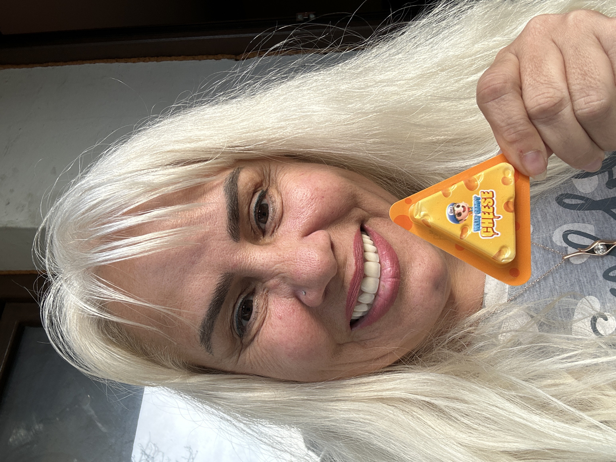 A woman is shown in a close-up selfie, holding a small, triangular package of "Willy Buddy Cheese". The woman has blonde hair and fair skin, and she is smiling. She has a nose piercing on the left side. She is wearing a gray shirt with white heart patterns and a silver necklace with a pendant. The background shows a building entrance and the exterior. The location is Sofia, Bulgaria.