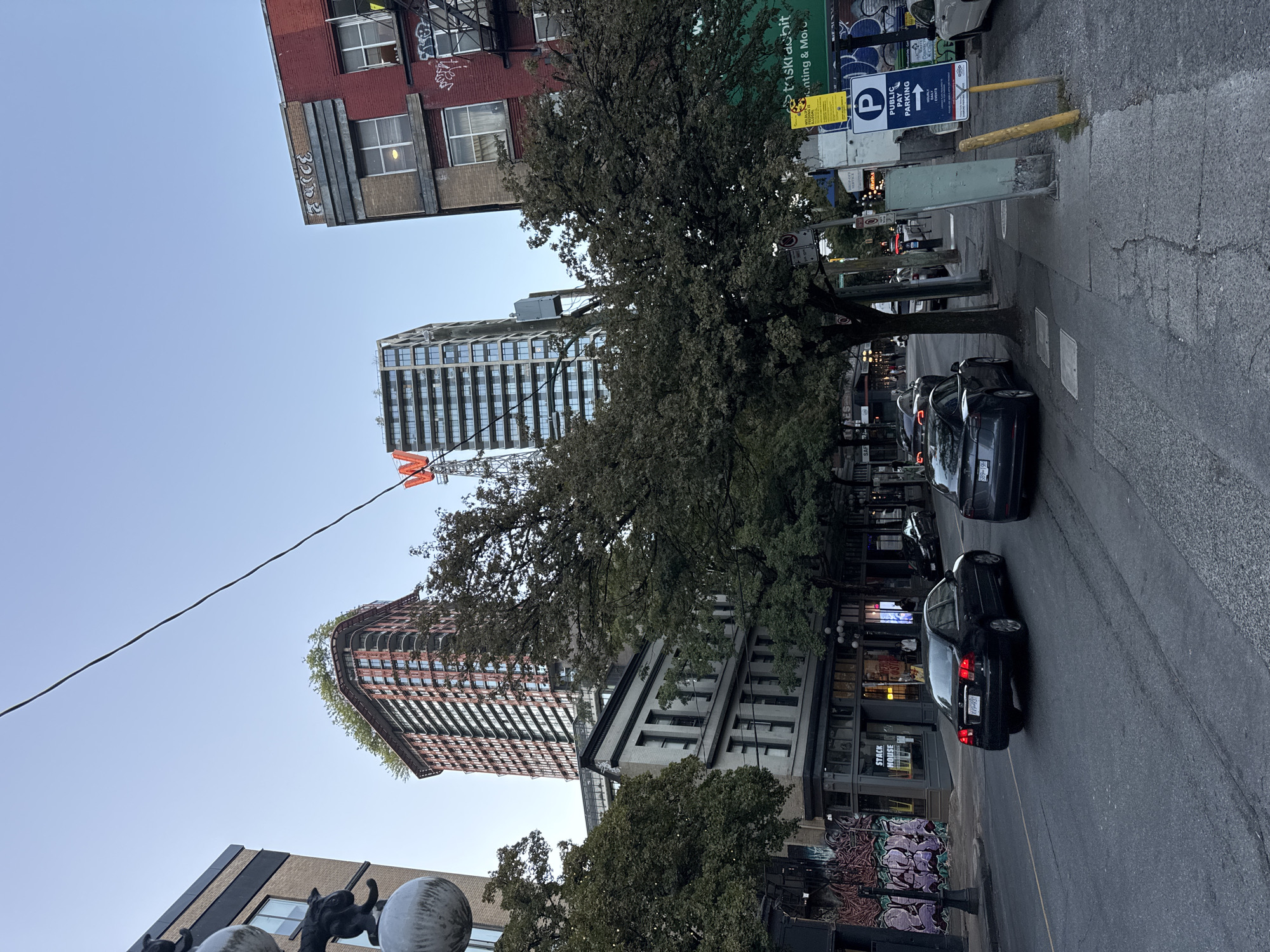 The image shows a street scene in Vancouver, Canada, likely in the Gastown or Chinatown area, judging by the architecture and signage. It is late afternoon or early evening, with the sky a clear pale blue. The street is paved with asphalt, showing tire marks.
In the foreground, two cars are visible on the street. The car closest to the viewer is a dark-colored sedan, possibly a Tesla, with its license plate partially obscured. Behind it is another dark sedan, a Honda Civic, with its taillights illuminated.
The sidewalk is made of concrete and appears somewhat cracked. Several trees with full green foliage line the sidewalk, providing shade. On the sidewalk, there are various street signs. One prominent sign indicates "PUBLIC PAY PARKING" with an arrow pointing to the right. Another sign, a yellow one with text and an image of a yellow jacket, appears to be a warning or informational notice. A light green utility box is also present near the sidewalk.
In the background, several buildings are visible. To the left, a brick building with a beige facade stands, adorned with graffiti on its lower sections. Next to it, a taller, modern building with a red and orange patterned facade rises, topped with a green roof of trees. Further to the right, a red brick building with multiple windows and fire escapes is prominent, also bearing graffiti. Adjacent to this, a tall, modern glass and concrete skyscraper stands, with a large orange "W" logo visible near its top, possibly indicating a business or landmark. Power lines crisscross the sky above the street.
There are no clear individuals in the image, though shadows suggest the presence of people in the background near the buildings. The overall impression is an urban environment with a mix of older and newer architecture, a common sight in Vancouver's historic districts.