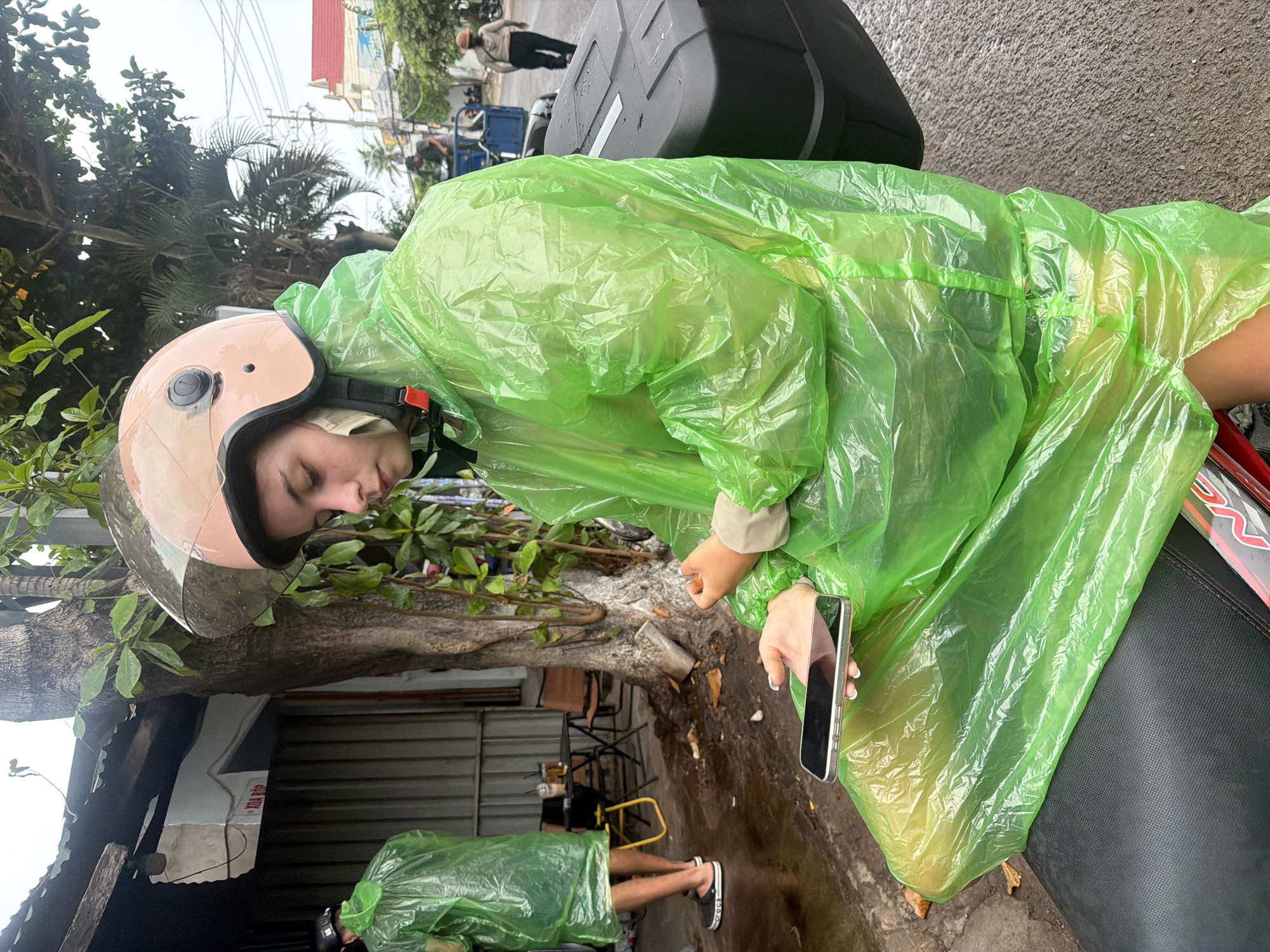 A young woman wearing a lime-green, crinkled plastic raincoat and a light pink helmet is sitting on a motorcycle. She is looking down at a smartphone in her hands. The phone is silver. Her raincoat is gathered at her wrists, which are pale. She has long, dark hair, and her face is partially obscured by the helmet visor. In the background, another person in a similar green raincoat stands on a wet, paved street. Behind them is a row of buildings, some with corrugated metal siding. Trees with dense foliage are visible, and the sky is overcast. The overall setting suggests a street scene in Nha Trang, Vietnam, possibly during a rain shower, given the wet surfaces and raincoats.
