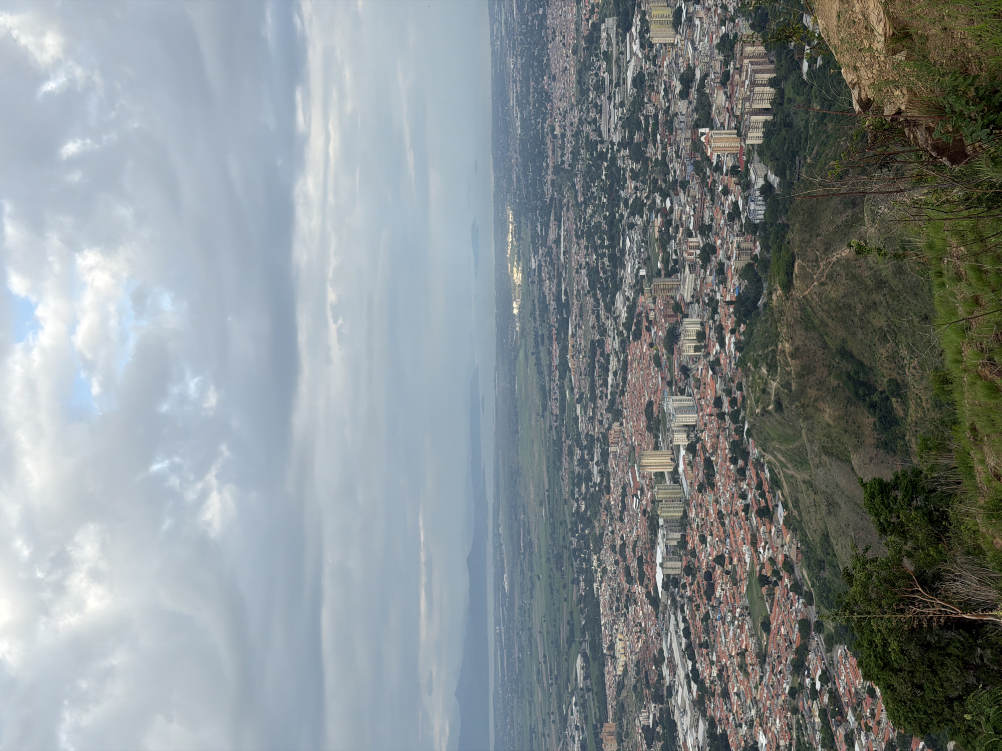 This is a high-angle, wide shot of a densely populated urban area in El Cambur, Venezuela, with a body of water and distant mountains in the background. The foreground is dominated by a steep, grassy slope with some dry brush and rocks. The midground shows a sprawling city with a mix of residential buildings, characterized by red-tiled roofs, and taller, more modern apartment buildings and commercial structures, some with balconies. The urban landscape extends to the horizon, where it meets a vast, calm body of water, possibly a lake or bay, with islands or peninsulas visible in the distance. Beyond the water, a hazy mountain range forms the backdrop. The sky is overcast with dramatic, textured clouds, suggesting a cloudy or potentially stormy day. There are no people or distinct objects clearly visible in the scene. The overall impression is one of an extensive, urbanized area situated within a natural landscape.