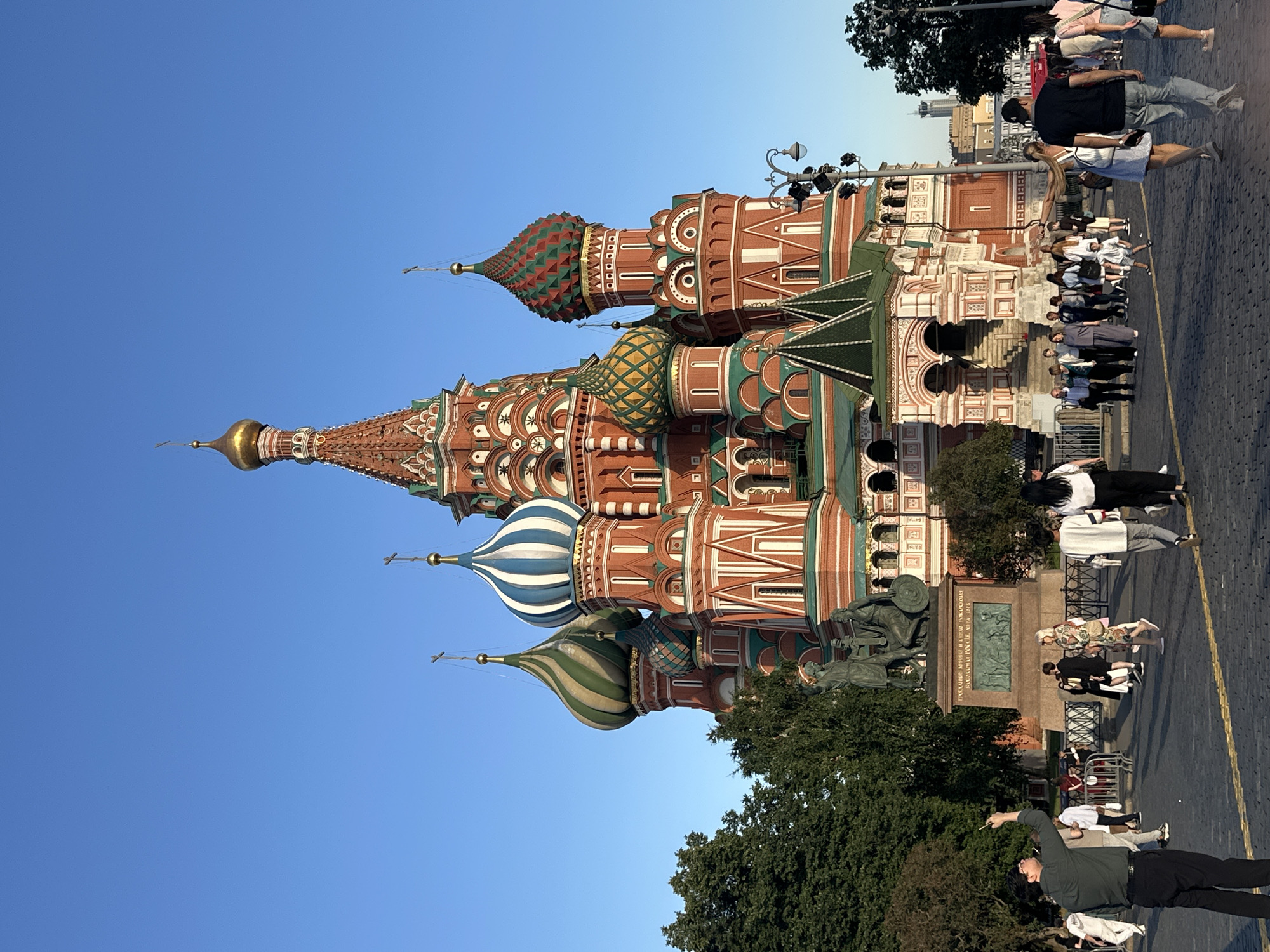 This is a full shot, eye-level, outdoor, daylight, photograph of Saint Basil's Cathedral in Moscow, Russia. The iconic cathedral, with its vibrant colors and distinctive onion domes, is the central subject, occupying the majority of the frame. The sky is a clear, bright blue. In the foreground, a paved plaza is visible, with various people walking around. A man in a dark green shirt and black pants is in the left foreground, facing away from the camera and holding a phone up, as if taking a picture. To his right, a bronze statue on a pedestal is situated. Beyond the statue and to the right of the cathedral, a lamppost with multiple lights stands. Trees are visible to the left of the cathedral, and other buildings and trees are in the background. The people in the scene are diverse in age and attire, with some dressed casually and others in more formal wear. Some individuals are walking in groups, while others are alone. The overall impression is that of a tourist destination on a sunny day.