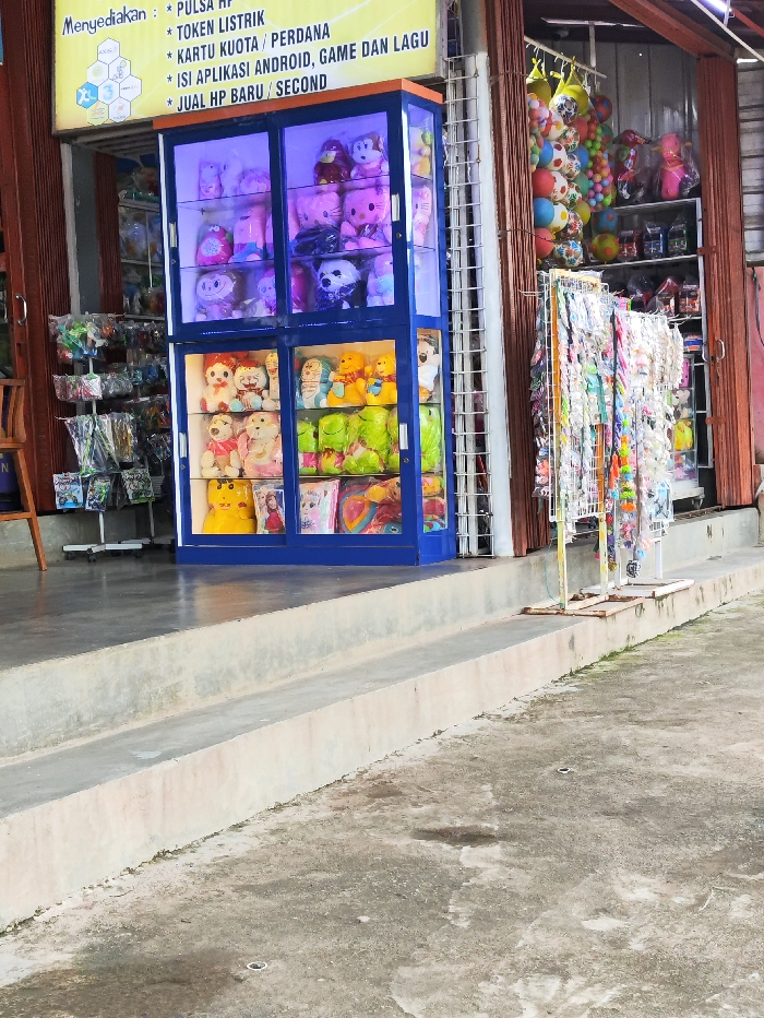 The image displays the exterior of a small shop in Selagai Lingga, Indonesia, during what appears to be daytime. The foreground shows a concrete ground with some stains and a few subtle indentations, leading up to two concrete steps that form a small raised platform in front of the shop.
The main subject is a prominent blue display cabinet, possibly for prizes or merchandise, filled with a wide assortment of colorful stuffed animals. The top section of the cabinet, illuminated with a purple light, showcases various plush toys, including teddy bears, a Hello Kitty figure, and other popular cartoon characters. The lower sections also hold numerous soft toys, such as Doraemon, more bears, and other vibrant figures.
To the right of this cabinet, a white metal stand displays multiple rows of hanging merchandise, which appear to be small toys, keychains, or accessories. Further inside the shop, shelves are visible, stacked with items like colorful inflatable balls and other packaged goods. The shop entrance features a brown, corrugated metal roll-up shutter, partially open on the right.
Above the blue cabinet, a yellow sign with black text advertises the services offered. The visible text reads: "Menyediakan: PULSA," "* TOKEN LISTRIK," "* KARTU KUOTA / PERDANA," "* ISI APLIKASI ANDROID, GAME DAN LAGU," and "* JUAL HP BARU / SECOND." This indicates the shop offers mobile top-ups, electricity tokens, internet data cards, app/game/music installations, and sells new and used mobile phones. No people are visible in the scene.