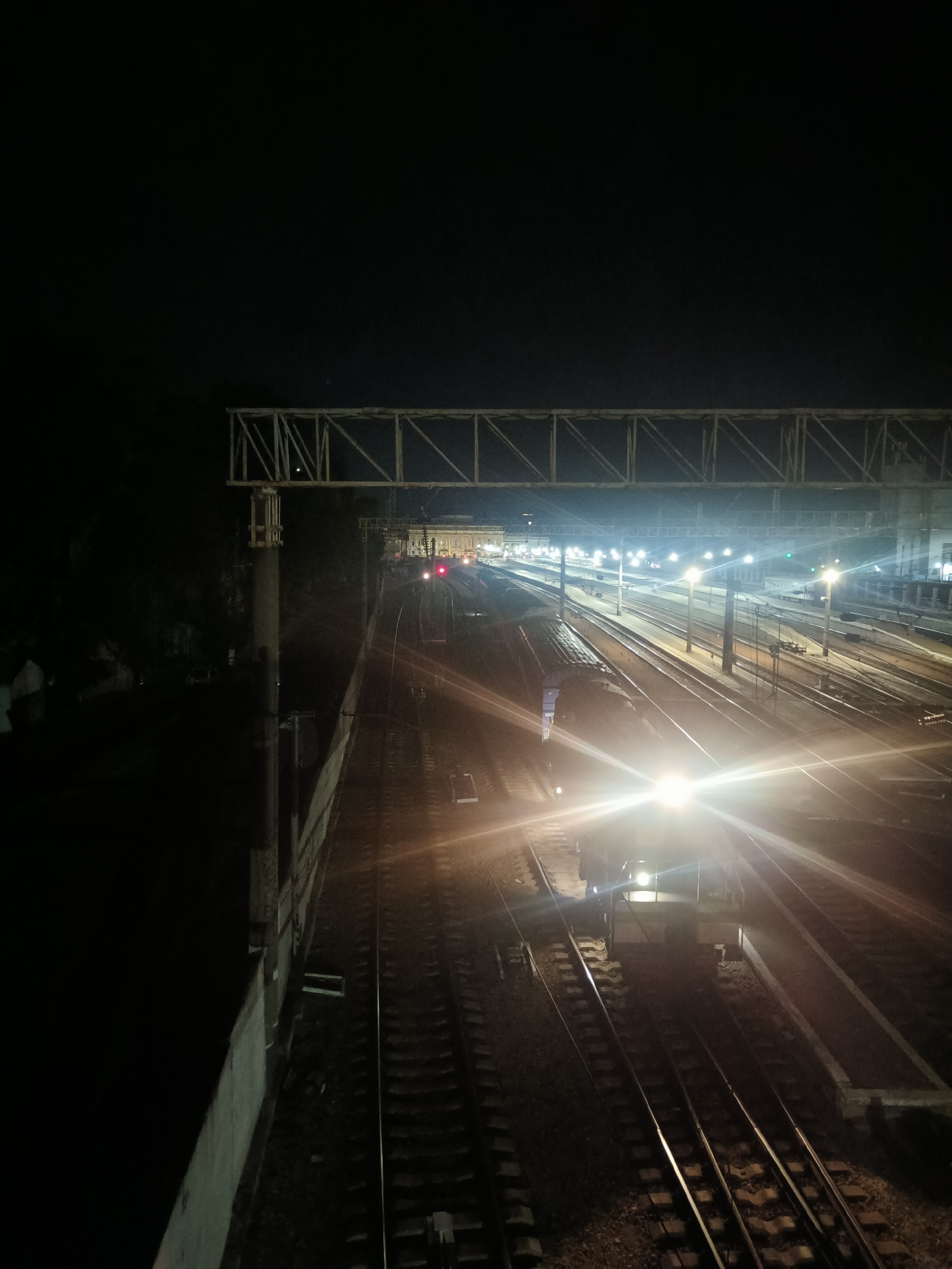 The image depicts a nighttime scene at a railway station, likely in Odesa, Ukraine. The perspective is from an elevated position, looking down onto multiple sets of train tracks. A train with its headlights on is visible in the foreground, casting a bright glare that obscures details of the locomotive. The station is illuminated by numerous lights, including streetlights and lights from buildings in the background, creating long streaks and reflections on the wet or polished surfaces of the tracks. Overhead structures, possibly gantries or bridges, span across the tracks. The overall environment is dark, with the artificial lights creating a stark contrast. There are no discernible people in the image, and the scene appears to be functional and operational, as indicated by the illuminated train and station lights.