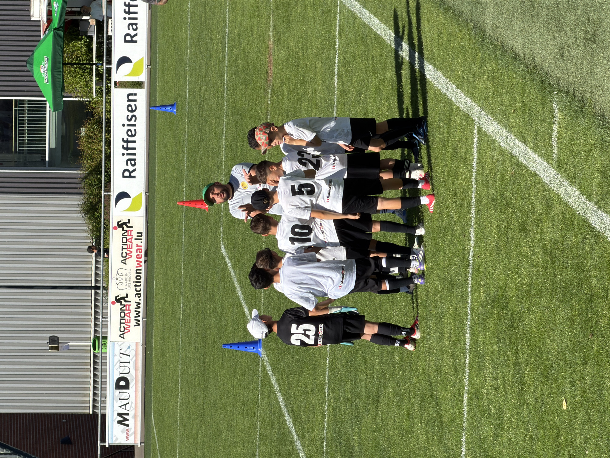 The image shows a soccer team of young players, likely in Fentange, Luxembourg, based on visible advertising signage. They are lined up on a grass soccer field along a white line, with a coach standing with them. The players are wearing white jerseys with black shorts, numbered 25, 10, 5, and 27. The coach wears a grey shirt and a green hat and is giving a thumbs-up gesture. There are orange and blue cones positioned at either end of the team. In the background, there are advertising signs for "MAUDUITZ", "ACTION WEAR", and "Raiffeisen", as well as a "BOFFERDING" umbrella. A building with corrugated siding is visible on the left side of the image. The lighting indicates a sunny day.