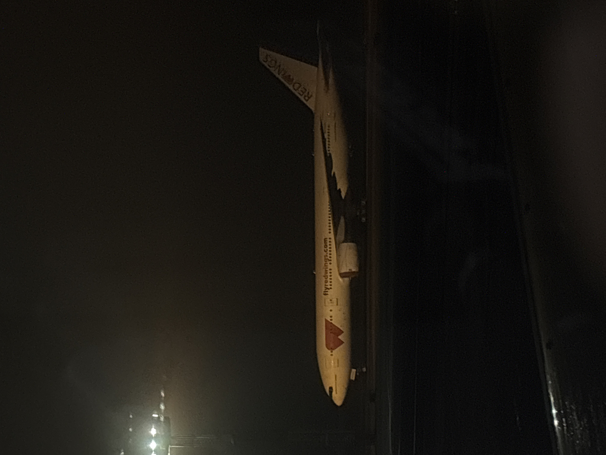 The image depicts the fuselage of a Red Wings airline plane. The location is unknown. The image is dark, making it difficult to discern details.