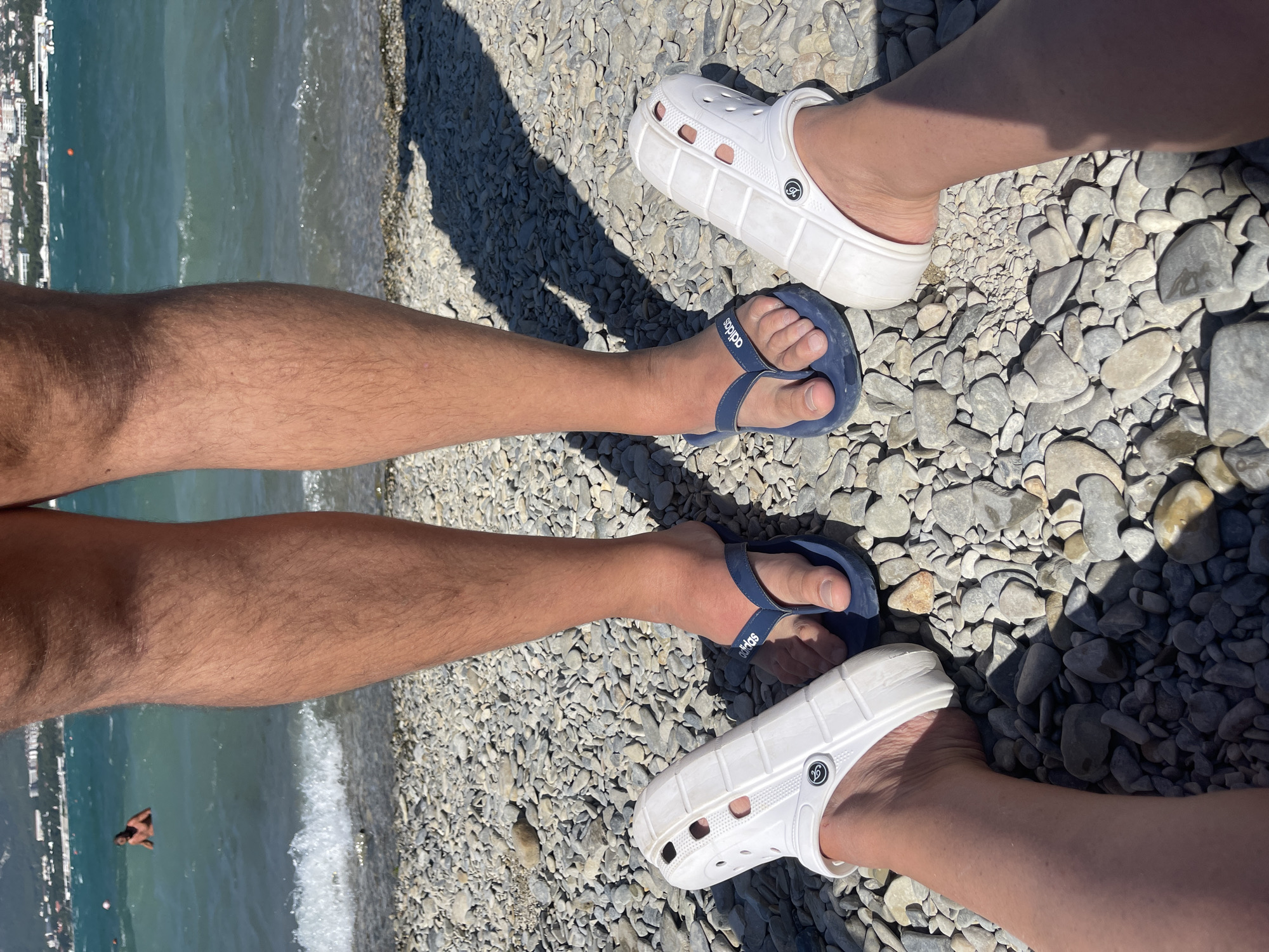 The image shows two pairs of legs with feet wearing footwear, standing on a pebble beach. In the background, the sea is visible, with a person swimming in the water and a cityscape on the distant shore, suggesting the location is Gelendzhik, Russia. One pair of feet is wearing blue flip-flops with the Adidas logo. The other pair of feet is wearing white Crocs. The legs are tanned and hairy, indicating a sunny outdoor environment. The pebbles on the beach are of various sizes and shades of grey and brown. The water is turquoise and appears to be choppy, with small waves breaking on the shore. The sky is bright and sunny, casting shadows on the beach. The cityscape in the background consists of buildings and trees.