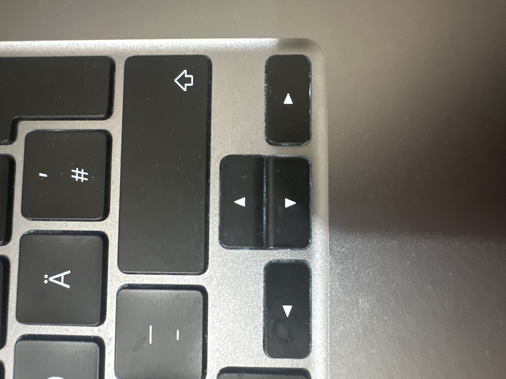 The image shows a close-up view of a portion of a laptop keyboard. The keyboard is a dark color with silver or gray lettering. Several keys are visible, including a key with the letter "Ä" and two dots above it, a key with a single apostrophe and a hash symbol (#) below it, a key with two horizontal lines, a large rectangular key with a symbol that resembles an upward-pointing arrow, and a set of arrow keys: left, up, down, and right. The keys appear to be slightly worn. The surrounding area of the keyboard is also visible, made of a metallic material. The image is taken from a top-down perspective, and there is no visible indication of the city of Hamburg, Germany, or any other geographical location.