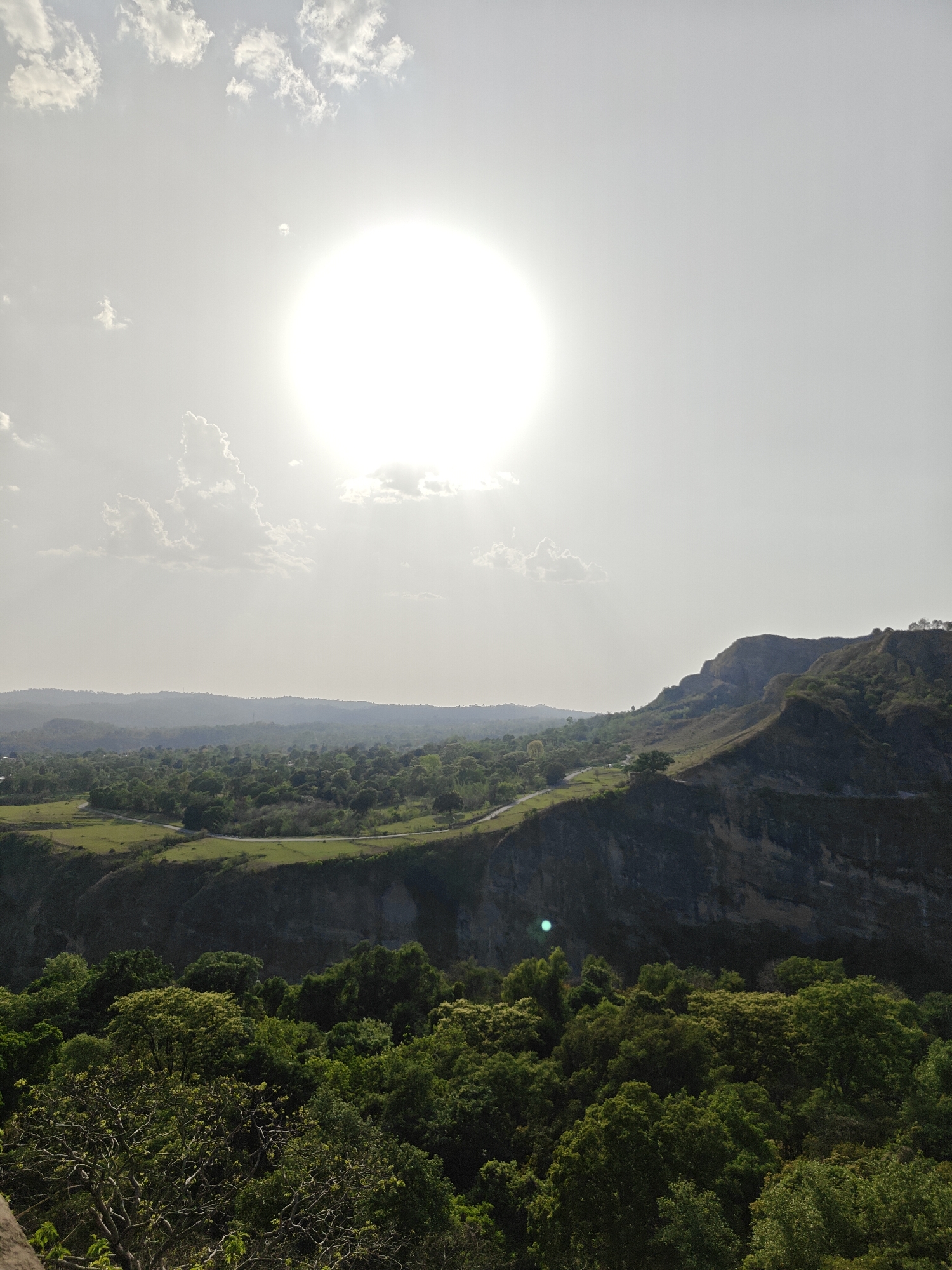 This image depicts a landscape in Kaswara, India, under a bright, overcast sky. The dominant feature is a series of rolling hills in the midground, covered in sparse vegetation and leading to a taller, more rugged mountain ridge. A road winds along the hillside. In the foreground, there's a dense forest canopy, with individual trees distinguishable by their varying shades of green. A distinct, though less prominent, valley stretches beyond the hills in the midground, dotted with agricultural fields and patches of trees. The sun appears as a large, diffused orb in the sky.
