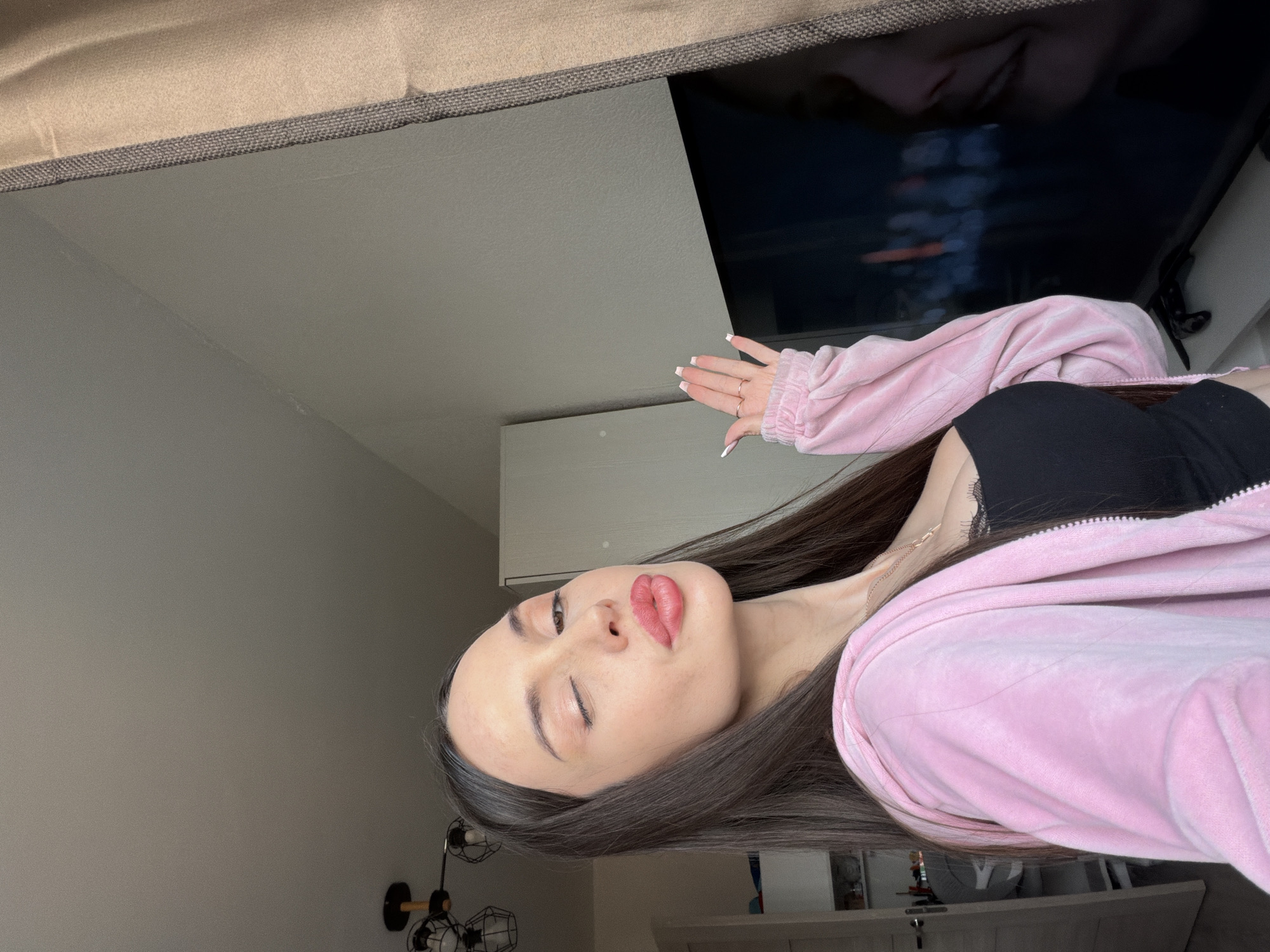 A young woman with long, dark hair is photographed in a domestic interior. She is wearing a black top and a pink velour jacket. She has fair skin and prominent lip makeup. One eye is closed, suggesting a wink or a pose.
The background includes a television screen, a white cabinet, and a lamp with multiple light fixtures. The presence of a television, cabinet, and lamp suggests this is a living room or bedroom.
There is no evidence to support that this image was taken in the city of Saint Petersburg, Russia.