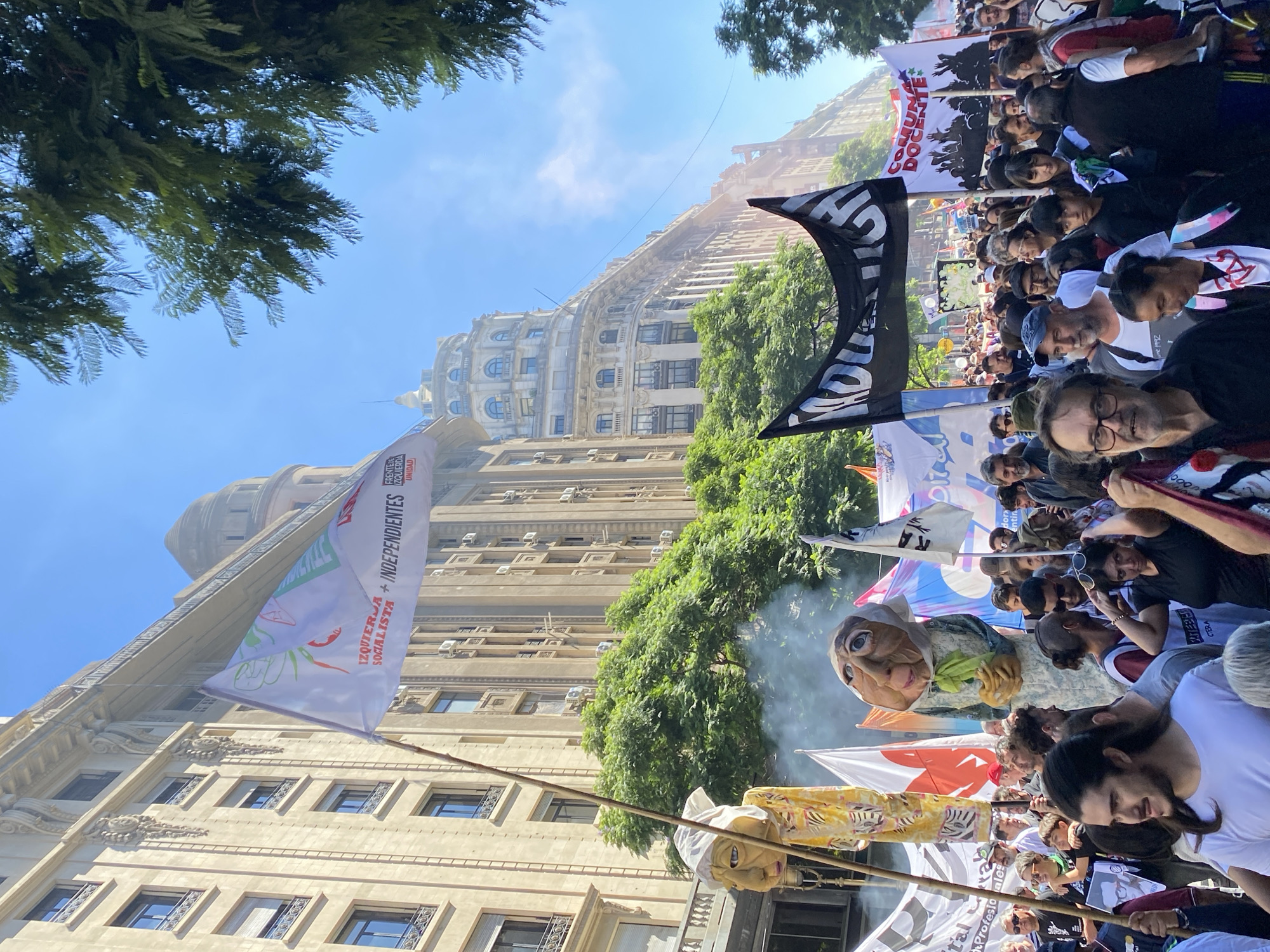 The image depicts a crowded street protest under a clear blue sky on **Avenida de Mayo in Buenos Aires, Argentina**. Tall, ornate multi-story buildings with European architectural influences line both sides of the street, featuring numerous windows, balconies, and air conditioning units. Green trees with dense foliage are interspersed along the avenue.
A large number of people occupy the street, appearing to move forward in a march. Individuals are diverse in age and gender, dressed in casual attire. Many participants carry flags and banners. Prominent among these is a white flag held aloft on a bamboo pole, displaying red text that reads "IZQUIERDA + INDEPENDIENTES" (Left + Independents) and partially visible "FRENTE DE IZQUIERDA UNIDAD" (Left Front Unity). Another large black banner with white lettering, partially obscured, is visible further back. A white banner with black silhouettes of raised fists and the text "COMUNA DOCENTE" (Teaching Commune) is also present.
Two large, stylized effigy puppets are held above the crowd. One on the left features a yellow and white head with a stylized face and cloth head covering. A larger puppet in the center depicts an older-looking figure with a wrinkled face, white head covering, and a light-colored dress, holding a green object. Smoke rises from the area around this central puppet. In the distance, a grand building with a distinct dome is visible, characteristic of the architecture along Avenida de Mayo.