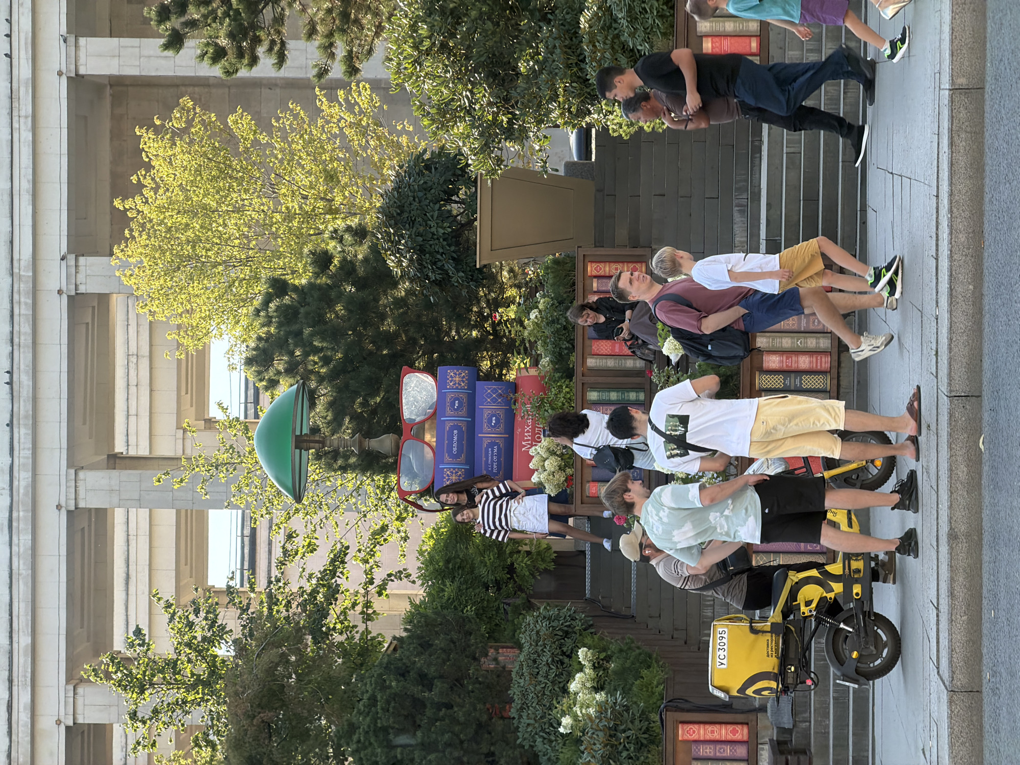 In an outdoor setting with a large building featuring a colonnade in the background, several people are visible on and around a set of wide stairs decorated with faux book spines. Two young females, one in a striped t-shirt and white skirt, the other in a black top, pose on the stairs. A group of individuals are gathered near the bottom of the stairs, including a man in a green t-shirt and black shorts, another man in a maroon shirt and blue shorts, and a young boy in yellow shorts. A yellow electric scooter with a delivery box is parked on the sidewalk in the foreground. Other people are walking past or observing the scene. The environment is landscaped with trees and bushes, and the scene appears to be a public space, possibly a plaza or park.