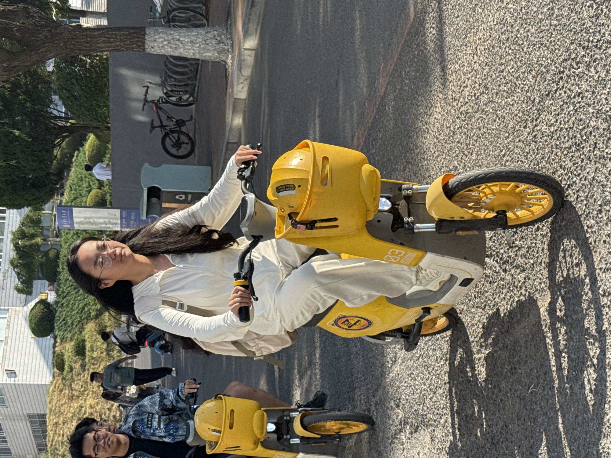 In Zhongtun, China, a young woman with dark, long hair and glasses is riding a yellow electric scooter. She is wearing a white jumpsuit and a light-colored backpack. Her left hand is on the handlebars, and her right hand is not visible. Another person is riding a similar yellow scooter behind her, to the left, and appears to be a young man with glasses, wearing a camouflage-patterned jacket over a dark shirt. In the background, there are trees, a building, and a bicycle rack with several bicycles. The ground is paved and textured. The lighting suggests it is daytime.