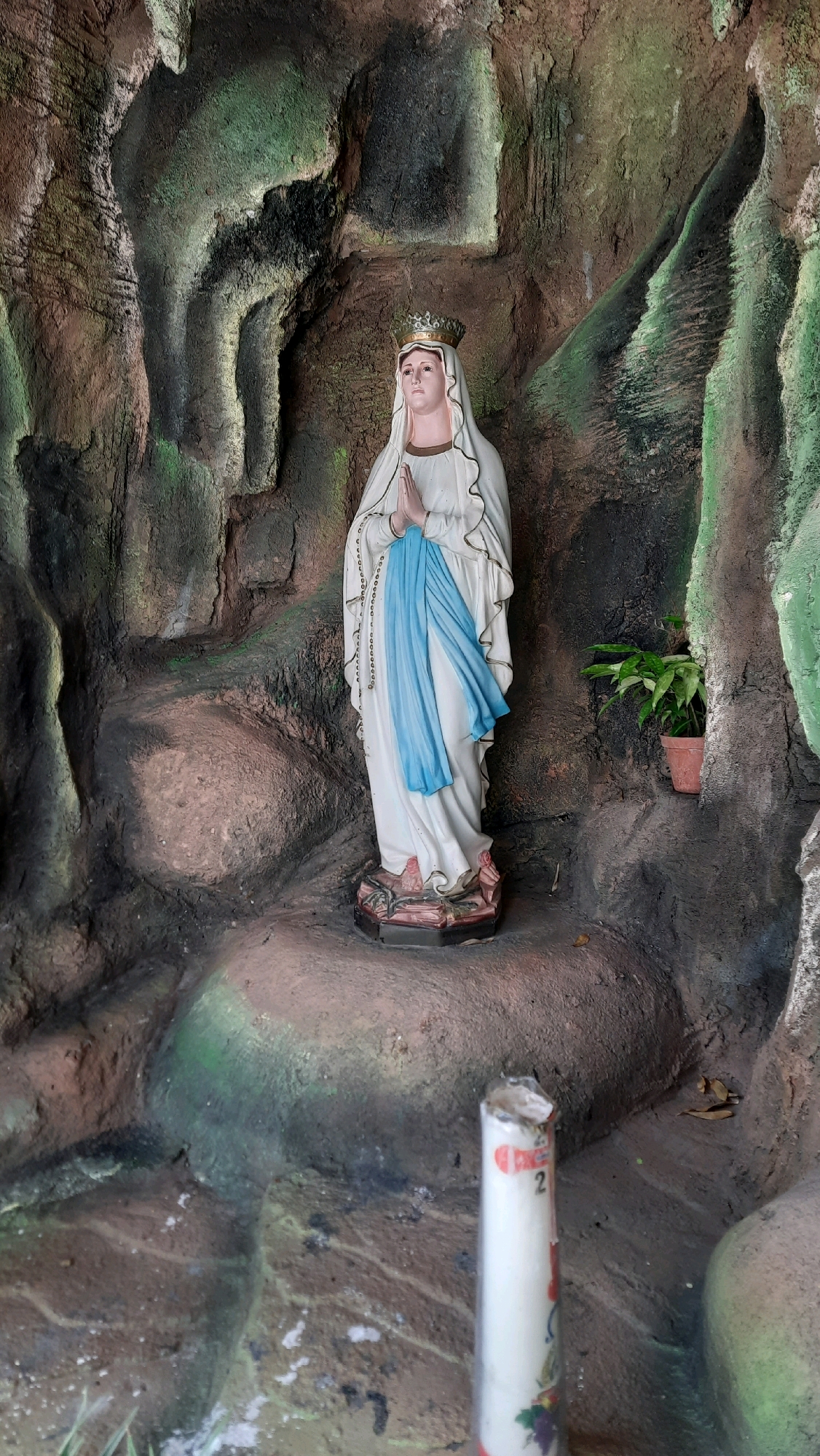 A statue of the Virgin Mary stands in a grotto. The statue depicts her with hands clasped in prayer, wearing a white robe, blue sash, and a crown. The grotto is made of rough, textured material resembling rock formations, with green mossy patches. To the right of the statue, a potted plant with green leaves sits on a ledge. In the foreground, a white candle with some red and green markings is visible. The scene is set within the city of Nanggulan, Indonesia.