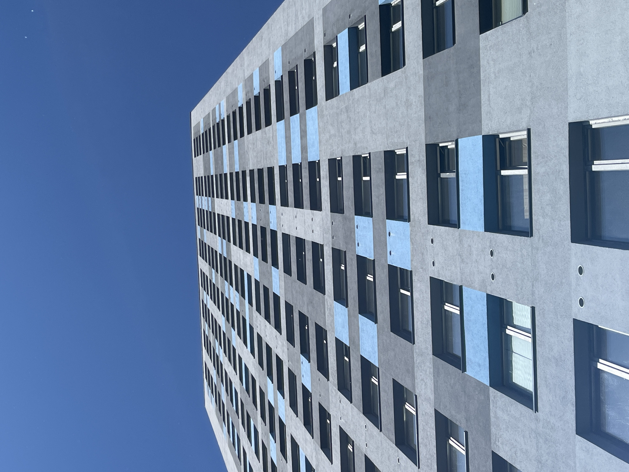 This media file is a low-angle, close-up shot of a modern building with a textured grey facade and alternating blue panels. The building has numerous dark-framed windows, some of which reflect the bright blue sky. The architecture suggests a contemporary urban setting. The shot is taken from a perspective looking upwards, emphasizing the height and scale of the building against a clear, cloudless sky. The overall impression is one of clean lines and a structured environment. The lighting suggests it is daytime, with the sun illuminating the building. No people or specific activities are visible, the focus is solely on the architectural elements. There is no visible text in the image.