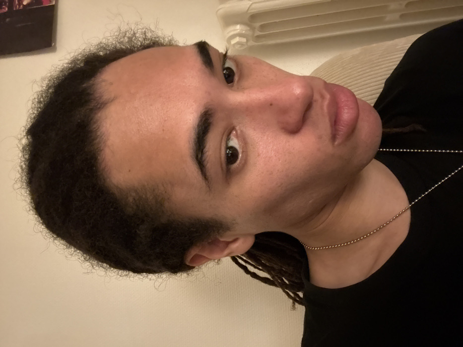 A person with dreadlocks is facing the camera. They have dark hair and light skin. Their eyebrows are thick and arched. They're wearing a black shirt and a silver beaded necklace. The background is a light neutral tone. A white radiator is partially visible behind their head.