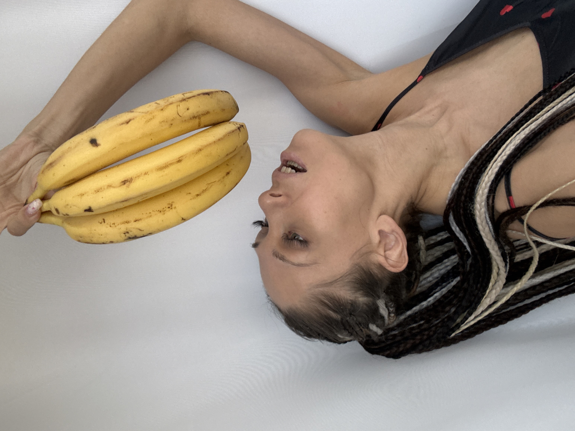 A light-skinned woman with braided hair lies on a white surface. She wears a dark top with red heart patterns. Her right arm extends upward, holding a bunch of four bananas. The photograph appears to have been taken from an elevated angle, facing down on the woman and bananas. There is no clear indication of the location being in Rusanovo, Russia.