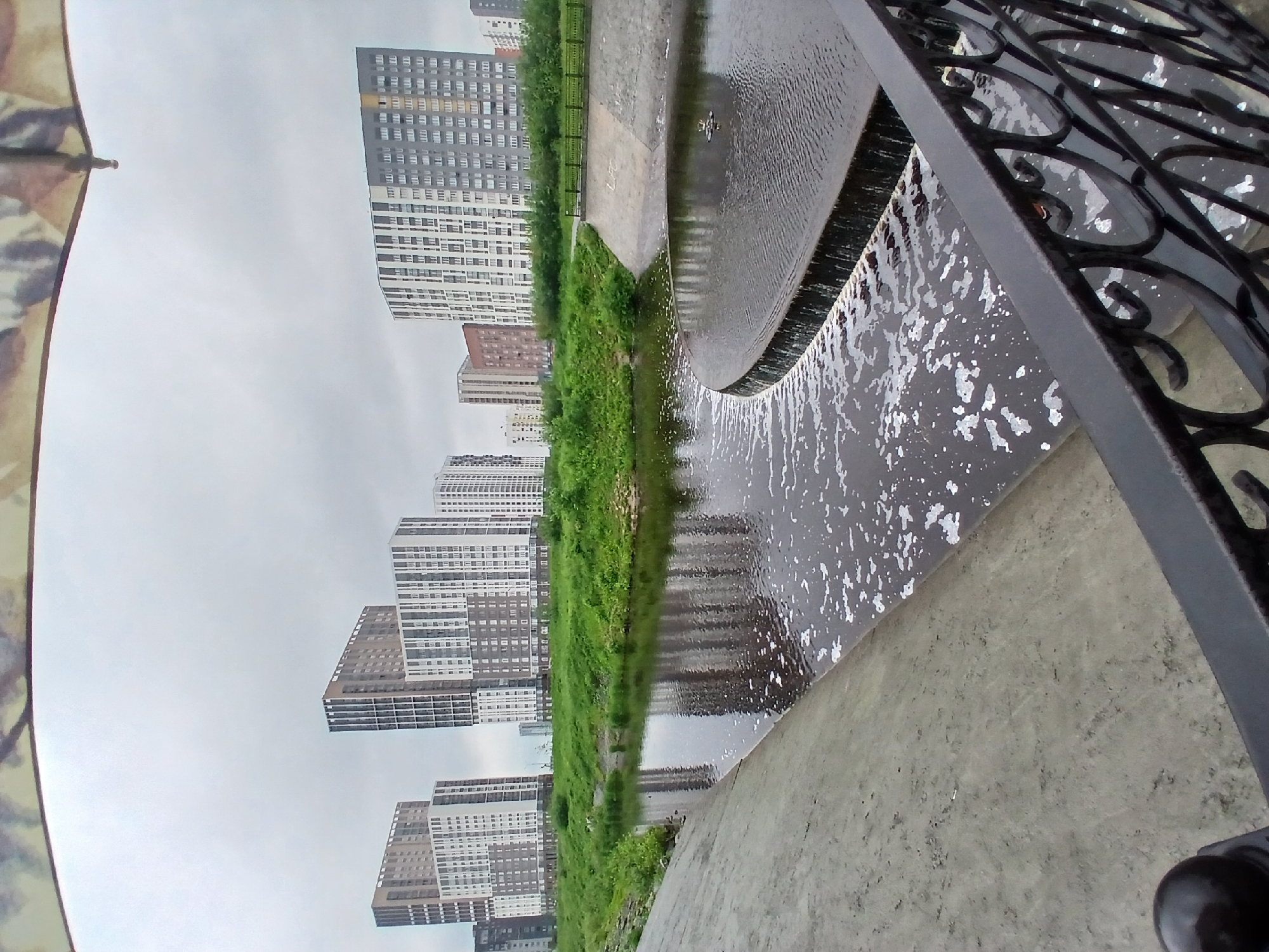 The image depicts a grey, overcast day in a city. In the foreground, a dark metal railing with an ornate design runs diagonally across the bottom of the frame. To the left, a concrete embankment slopes down towards a body of water. The water, reflecting the cloudy sky, has white foam scattered across its surface. A curved weir creates a small waterfall to the right of the frame, with more foam appearing below it. Beyond the water, a strip of lush green grass and trees separates the embankment from a line of modern, high-rise apartment buildings. These buildings are diverse in their architectural styles and colors, ranging from white and grey to brown and beige, with many visible windows. The buildings are characteristic of the residential areas developed in Yekaterinburg, Russia, particularly in newer districts. The overall scene suggests an urban park or waterfront area designed for residential living.