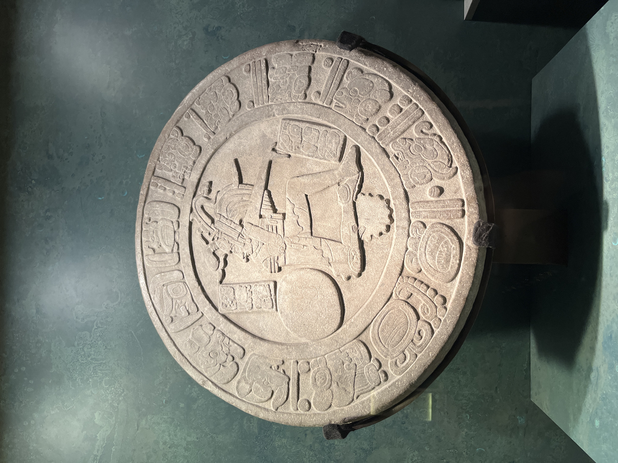 A circular stone artifact, possibly a relief carving or a calendar stone, is displayed on a dark stand against a mottled teal background. The artifact features intricate relief carvings that depict a central figure in a seated position, adorned with headdress and holding objects. The figure is surrounded by a ring of glyphs or symbols in a Mesoamerican script. The artifact appears to be made of light-colored stone, possibly limestone or sandstone, and exhibits signs of age and wear. The background wall has a textured, uneven surface with variations in color and light. The artifact is positioned in what appears to be a museum exhibit, with a dark shelf-like structure visible in the lower right corner casting a shadow. The overall lighting is diffused, highlighting the relief details of the artifact. This artifact is likely on display at the National Museum of Anthropology in Mexico City, Mexico, given the style and context of Mesoamerican artifacts typically housed there.