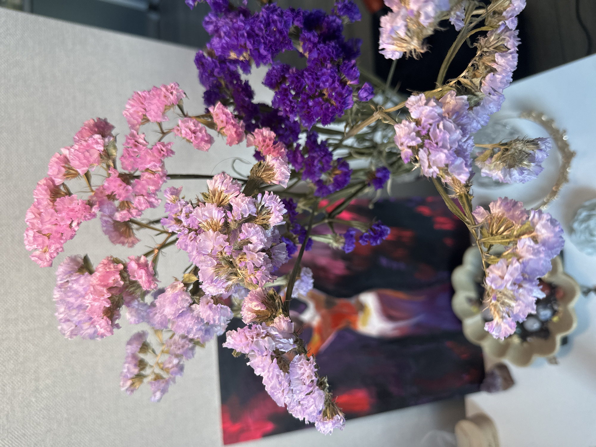 An indoor still life features a vase of dried flowers in shades of pink, purple, and lavender. The flowers are arranged in a bouquet, with some branches extending to the left and right. In the background, a painting with abstract figures in red and purple is visible. A white surface is below the flowers, with a decorative bowl and some small stones. The wall behind the flowers has a textured, light-colored finish. The scene is lit by an unseen light source, casting soft shadows.