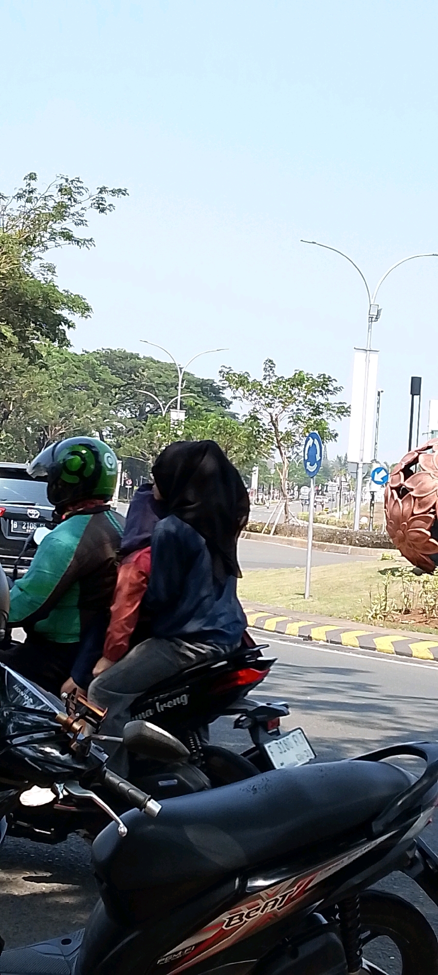 In Gunung Putri, Indonesia, on a sunny day, three individuals are riding on a single motorcycle. The rider in the front wears a green and black helmet, and a teal and black jacket. Seated behind the rider, a person in a pinkish-orange jacket and a dark blue head covering is partially visible. The third individual, seated furthest back, is wearing a black hijab and a dark blue long-sleeved shirt. The motorcycle, a Honda Beat, is parked on a paved road with yellow and black striped markings along the edge. In the background, trees, streetlights, and traffic signs are visible, suggesting an urban or suburban environment. The license plate of the motorcycle is partially obscured but appears to start with the letter "F".