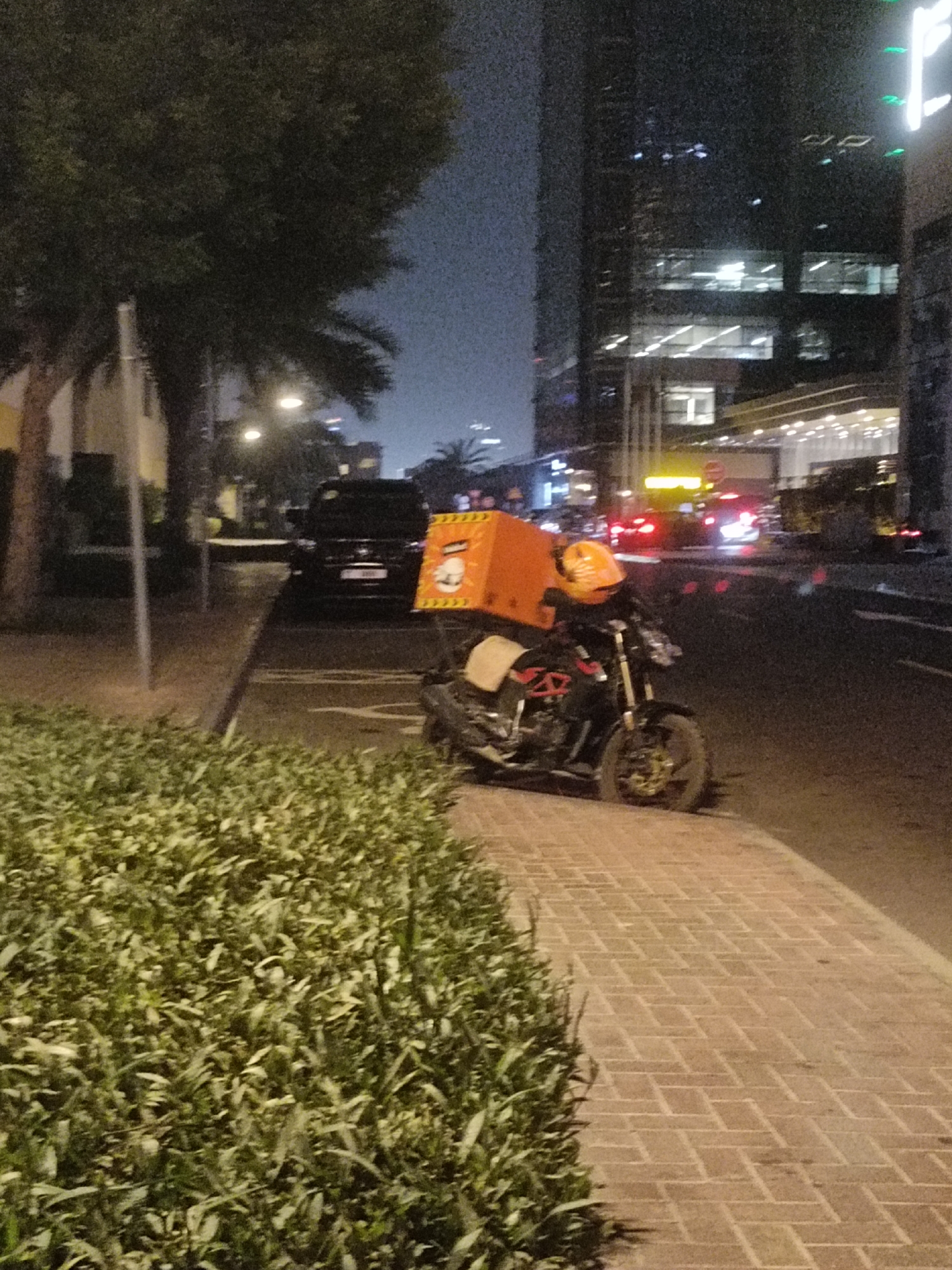 The image depicts a nighttime scene on a city street in Dubai, United Arab Emirates. In the foreground, a paved walkway made of brick is visible to the left, bordered by dense, green foliage. To the right of the walkway, a black motorcycle is parked. The motorcycle is equipped with a large, bright orange food delivery box on the rear and an orange helmet resting on the handlebars. The motorcycle is facing towards the right side of the frame.
Beyond the motorcycle, a black SUV is parked on the road. Further in the background, tall modern buildings with illuminated windows dominate the skyline, characteristic of Dubai's urban landscape. Streetlights are visible, casting a warm glow on the scene. The road surface is dark asphalt, and traffic lights and vehicle headlights can be seen in the distance, indicating ongoing activity. The overall atmosphere suggests a typical urban environment at night, likely in a commercial or residential district of Dubai.