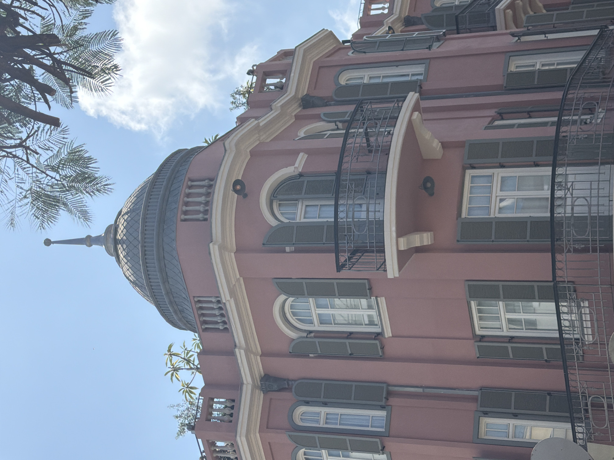 The image shows a building in Tel-Aviv, Israel. The building is multi-storied and has a dome-shaped roof. The facade is painted in a shade of pink with white trim. The windows are rectangular and arched with grey shutters. Some windows feature a metal balcony. Palm tree branches are visible in the upper left corner of the frame, set against a blue sky with sparse clouds.