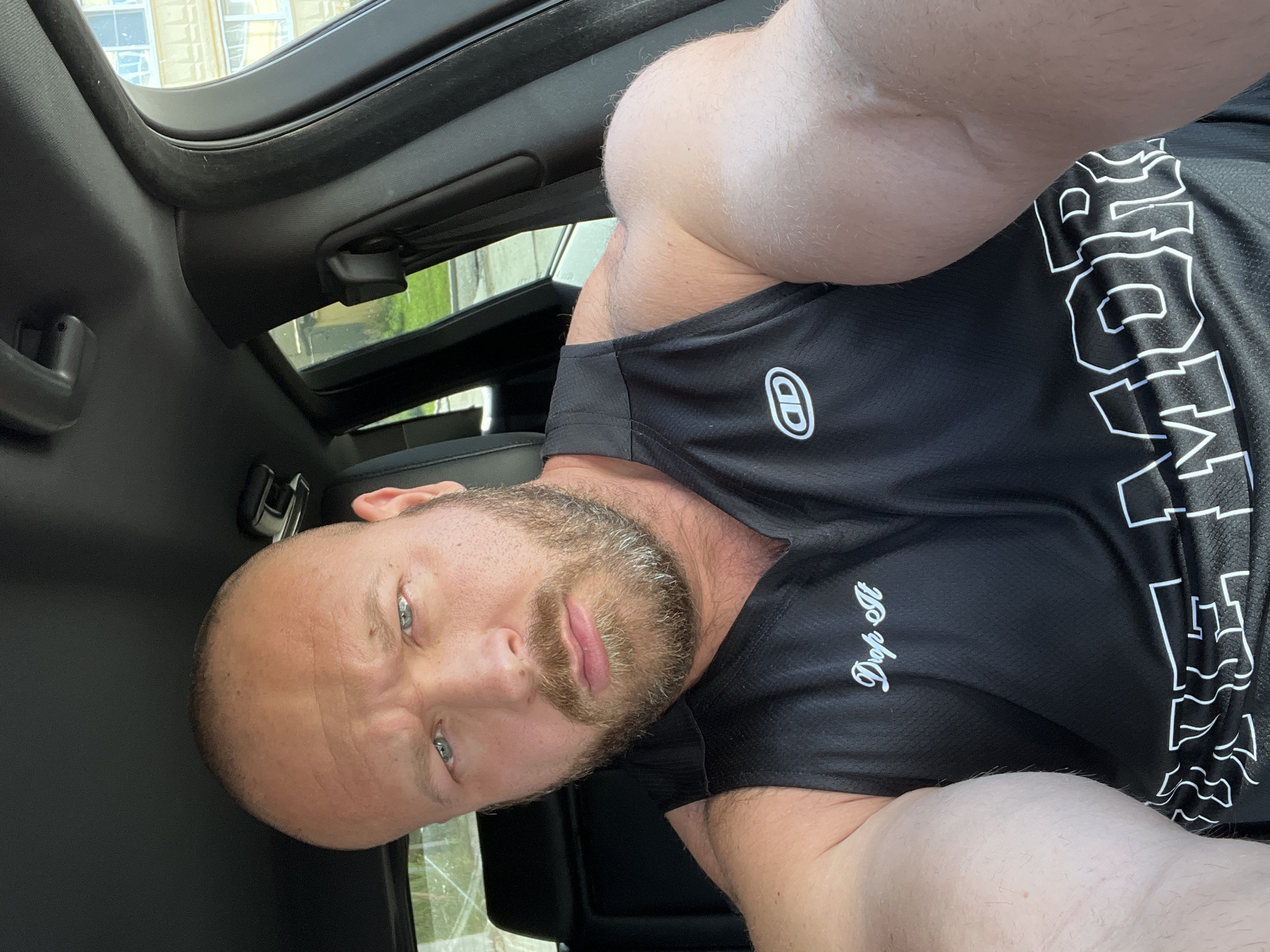 A bald, white man with blue eyes and a beard is sitting in the driver's seat of a car. He is wearing a black sleeveless shirt with white text and logos. His right arm is extended towards the camera, and his left arm is bent at the elbow. The car's interior is visible, including the dashboard and the ceiling. Outside the car, buildings and greenery are visible through the windows. The image appears to be taken in daylight. The man's expression is neutral.
