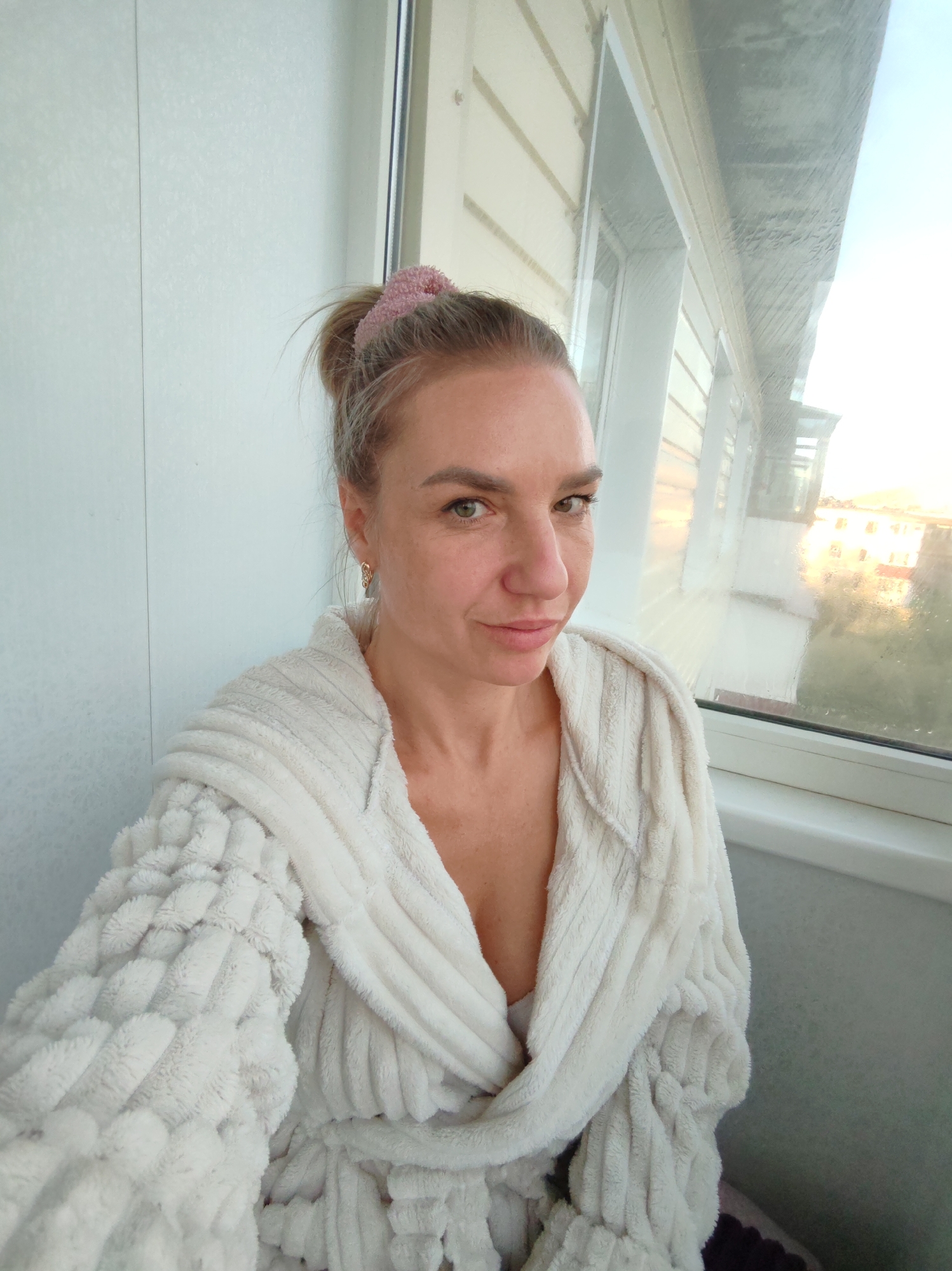A blonde woman with her hair in a bun tied with a pink scrunchie is pictured from the chest up. She is wearing a white fluffy robe with a textured pattern. Her left side faces the camera, and she is looking directly at the viewer. She has a gold earring in her visible ear. The background shows a window with condensation, and outside, buildings are visible in what appears to be a city environment. The lighting suggests it is either early morning or late afternoon. The overall scene appears to be an indoor residential setting, possibly a balcony or a room with a large window, in Petropavlovsk-Kamchatsky, Russia.