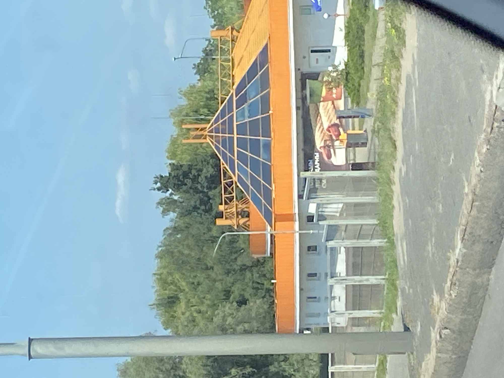 The image shows a modern building with an orange, pyramid-shaped glass roof and an orange trim. The building appears to be a commercial establishment, possibly a cafe or a restaurant, with a large advertisement on the side depicting food items. In front of the building, there is a grey concrete barrier, a paved area, and a fence. A tall grey pole, likely a lamppost, stands to the left of the frame. In the background, lush green trees line the horizon, and a clear blue sky is visible. The scene is set on a sunny day. This building is located on the Lenin Street in the city of Brest, Belarus.