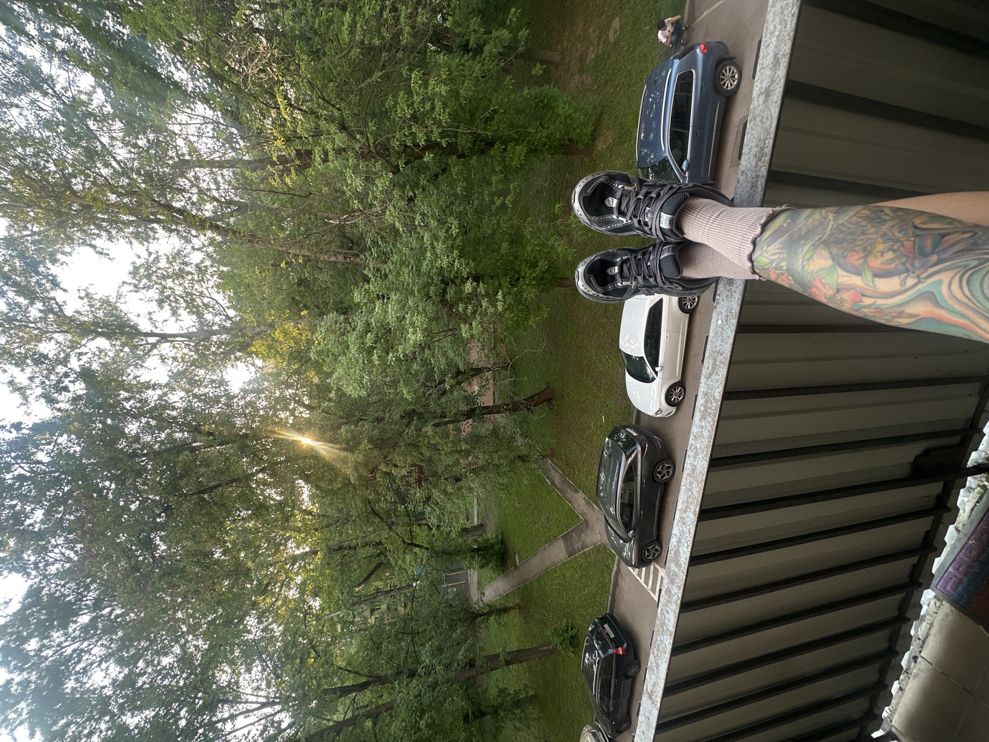 A downward-angle shot shows a person's legs, shod in gray athletic shoes and beige socks, extending over the edge of a balcony or elevated structure. A colorful tattoo adorns the visible forearm. Below, a parking area is visible with several cars of different colors, including white, gray, and dark blue. Further in the background, green trees fill the vista. Sunlight filters through the branches of the trees. The perspective suggests the photo was taken from an upper floor, providing a view of the outdoor environment below. The location is Moscow, Russia.