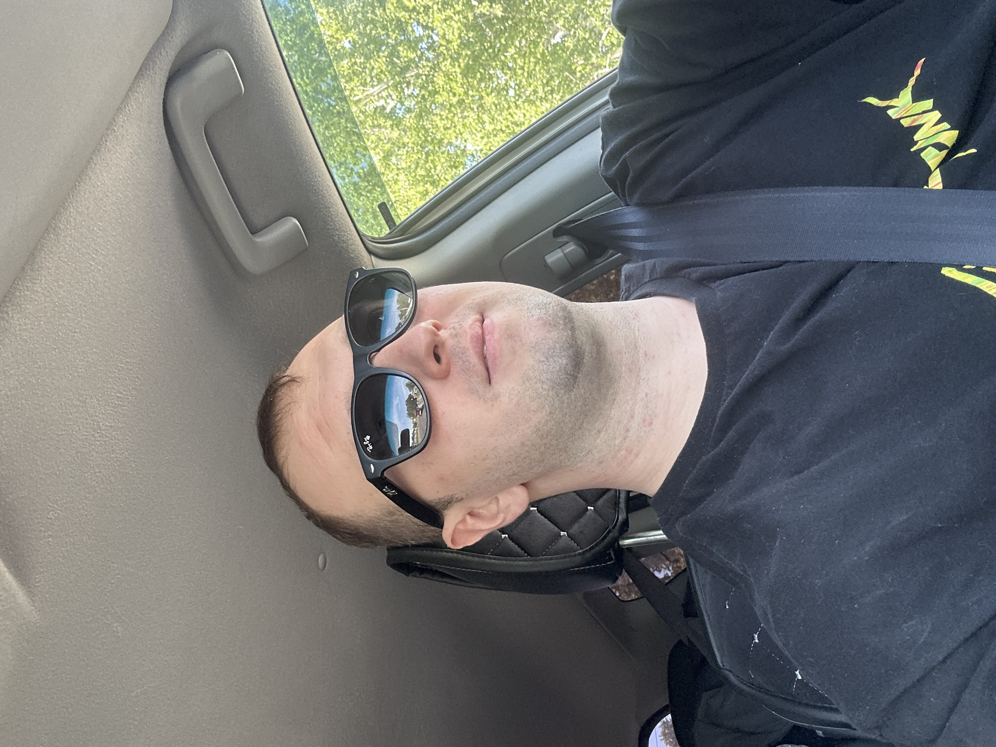 A man wearing a black T-shirt with a graphic design, sunglasses, and a seatbelt is inside a car. He has a bald head and stubble on his face. The interior of the car is visible, including the ceiling handle. Through the car window, trees are seen. It is impossible to determine the specific location in the city of Chaikovsky, Russia from the given image.