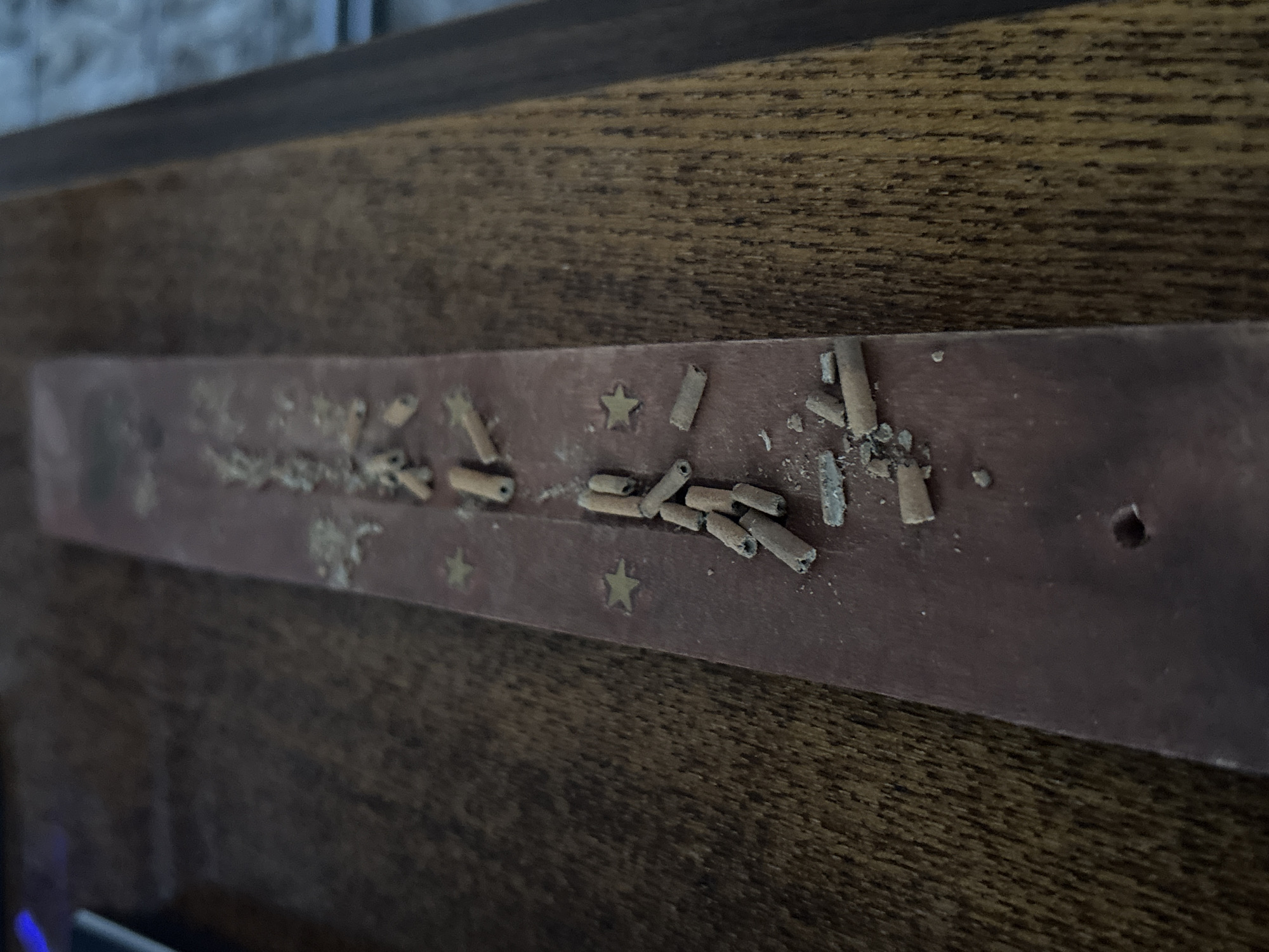The image shows a close-up, high-angle view of a dark reddish-brown, rectangular incense holder with small, cylindrical pieces of burnt incense scattered across its surface. The incense holder is decorated with small, yellow star shapes. The incense holder is placed on a dark, textured wooden surface. The scene does not provide any information that can be used to determine its location in Murmansk, Russia, or any details about people, actions, or interactions.