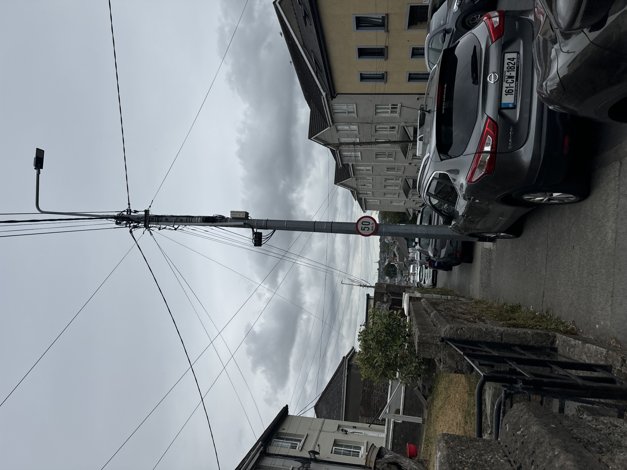 The image depicts a street scene in Waterford, Ireland, under an overcast sky. A gray Nissan Qashqai with license plate 161-CW-1824 is parked on the right side of the road. It is facing away from the camera and is positioned behind a black car. Further down the street, a row of white vans and other vehicles are visible. To the left, a row of houses with gray and yellow facades are seen. A utility pole with numerous wires runs vertically through the frame, and a circular speed limit sign indicating "50" is attached to it. The sidewalk is paved with concrete, and a stone wall with a black metal gate is located on the left side of the sidewalk. A lush green bush grows behind the wall. The overall impression is a residential street on a cloudy day.