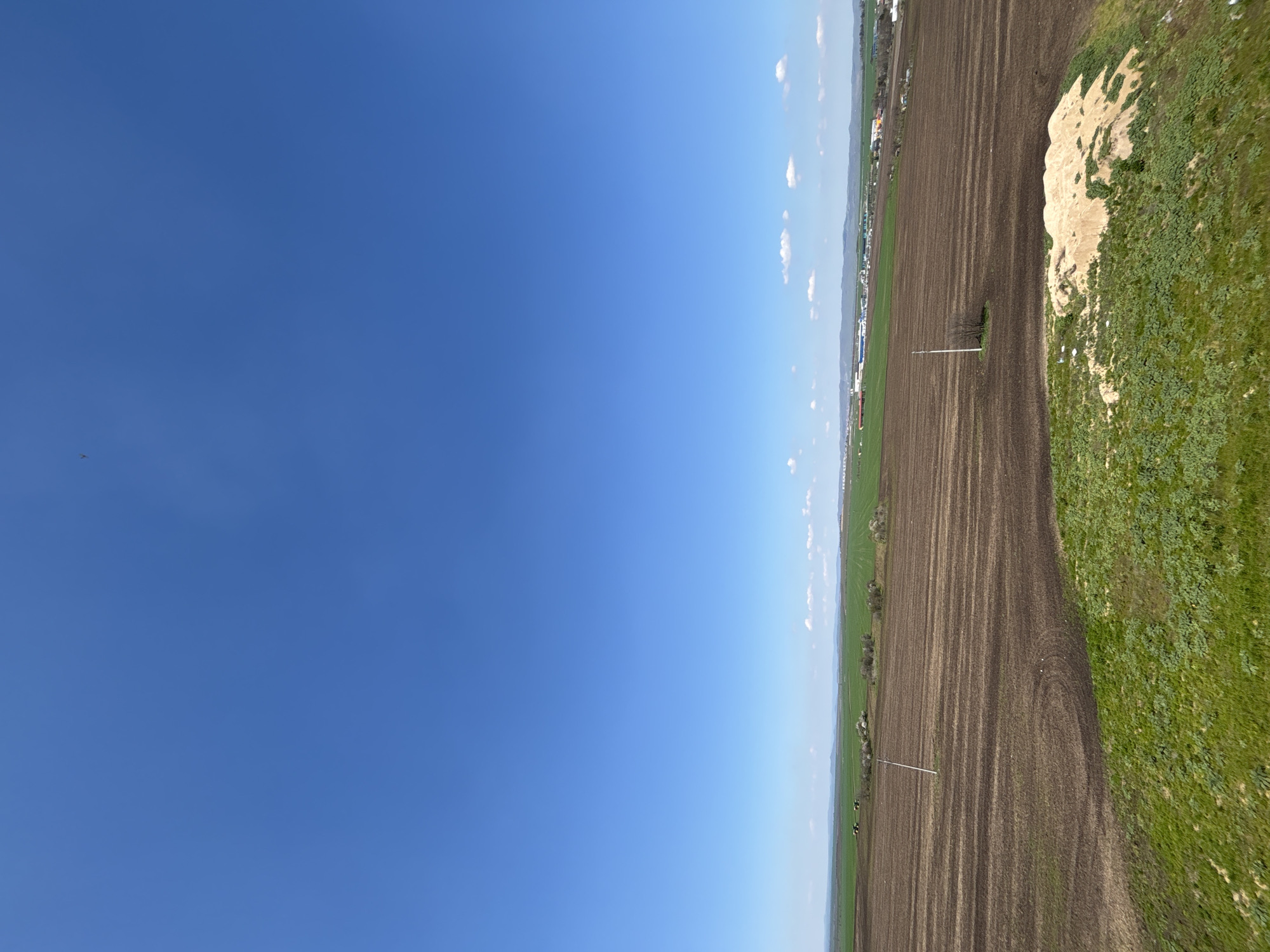 The image shows a landscape with a clear blue sky taking up about half the image. The other half features a view of terrain, including green vegetation, a dark brown field, and a strip of land that seems to have lighter-colored soil. Some sparse vegetation appears on top of this lighter area. There are some power poles situated on the edge of the dark brown field. In the distance is a stretch of land with buildings and hints of green spaces.
This is likely a rural area on the outskirts of Burgas, Bulgaria, based on the landscape characteristics, the presence of fields, and the type of vegetation. However, the image alone does not offer any information regarding Burgas.