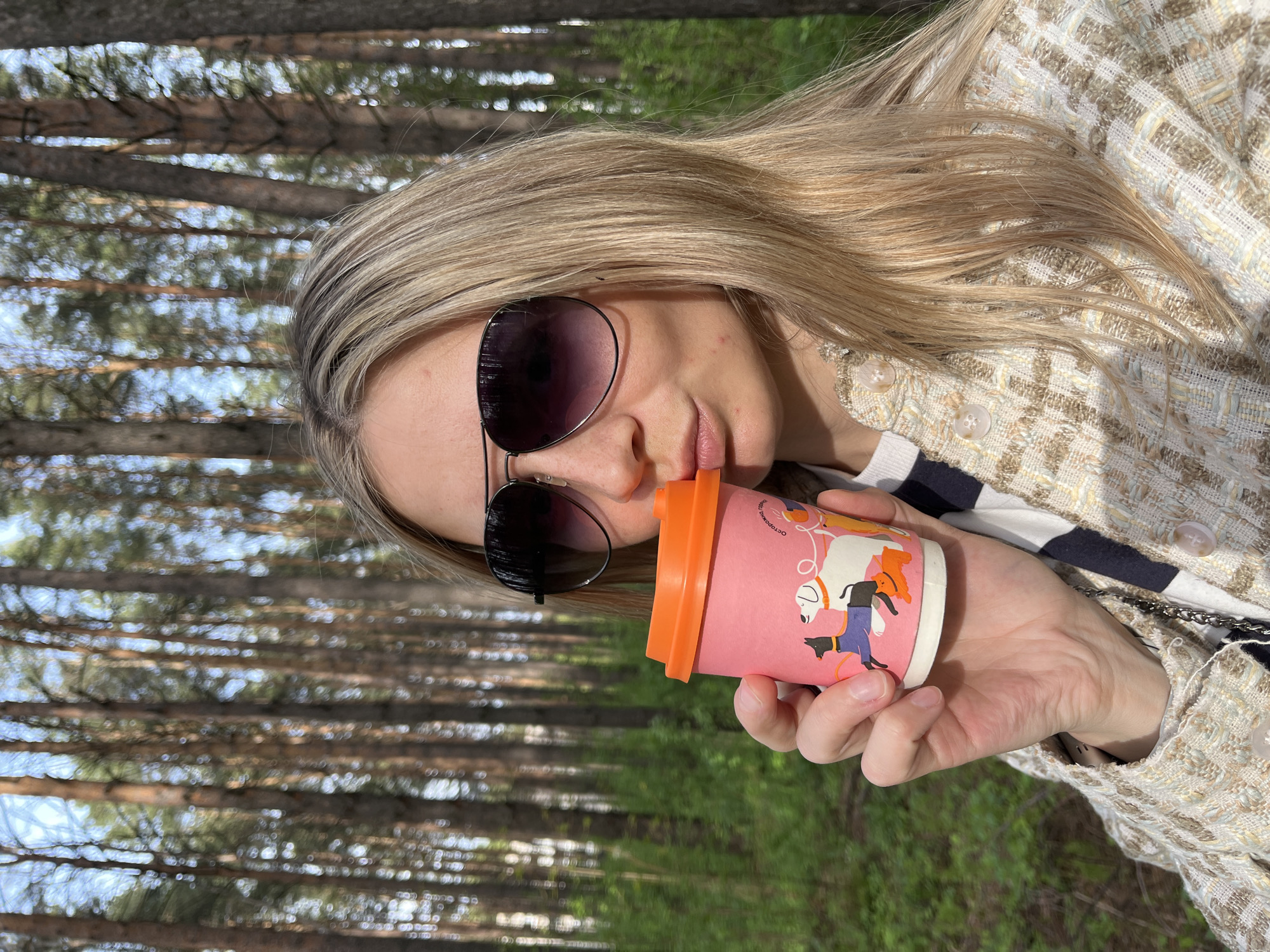 A woman is drinking from a pink cup with an orange lid in a forest, possibly on a sunny day. She is wearing sunglasses and a tan-and-white woven jacket over a striped shirt. The background consists of trees and green ground cover. The cup has a cartoon dog design on it. The location is Chaikovsky, Russia.