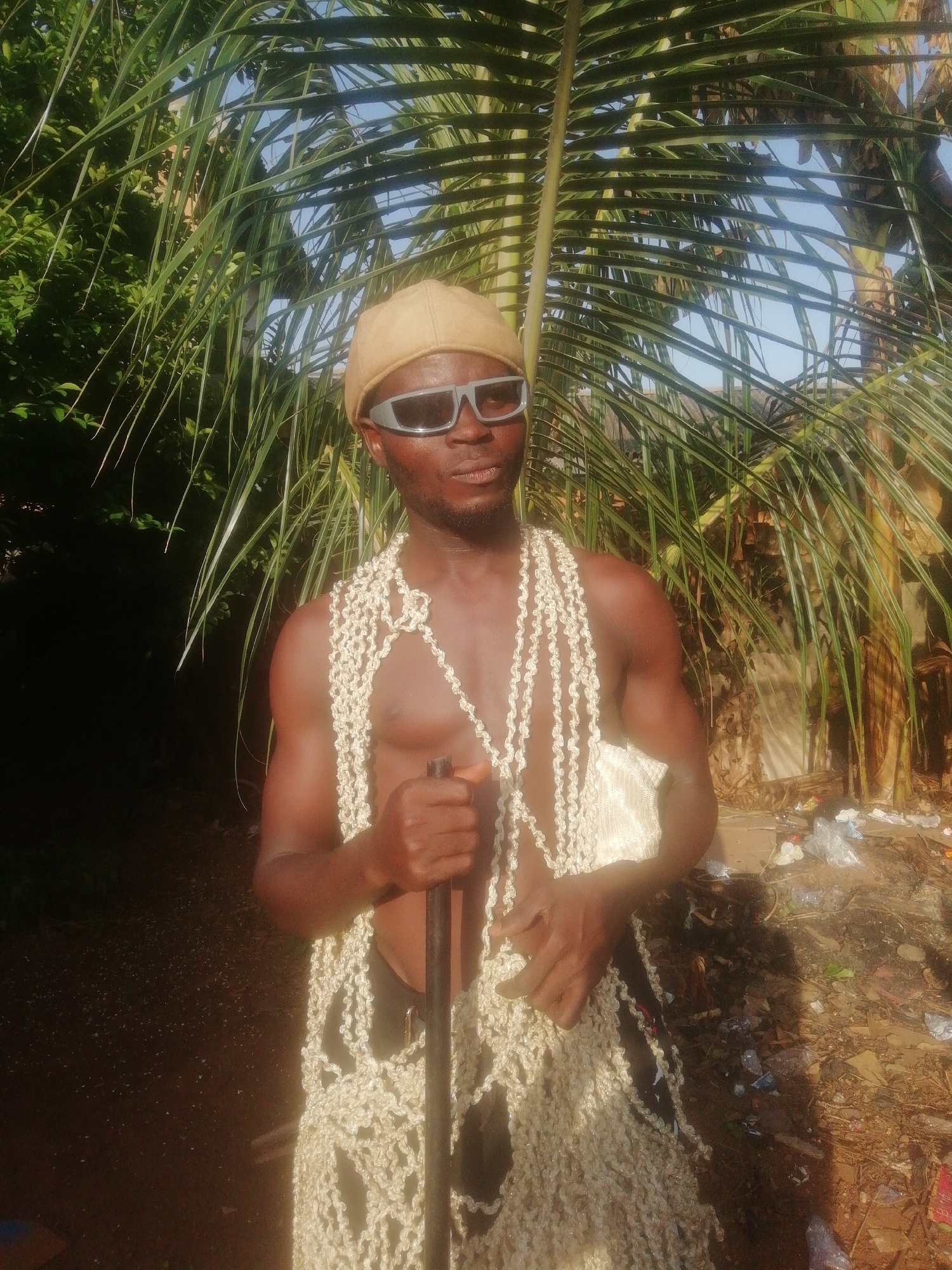 A medium shot captures a dark-skinned man with a beard, wearing a tan hat and silver sunglasses, standing in an outdoor setting, possibly in the city of Unknown. The man is shirtless, exposing his dark skin tone, and is adorned with what appears to be a garland or decorative net of white beads or seashells that covers much of his chest and torso. He firmly grips a dark, slender pole in his hands.
Behind the man, a palm tree with long, pointed fronds provides a backdrop, partially obscuring the sky. The area surrounding him appears unkempt with trash scattered on the ground, suggesting a neglected or potentially polluted environment. The lighting is bright, indicating it could be daytime.