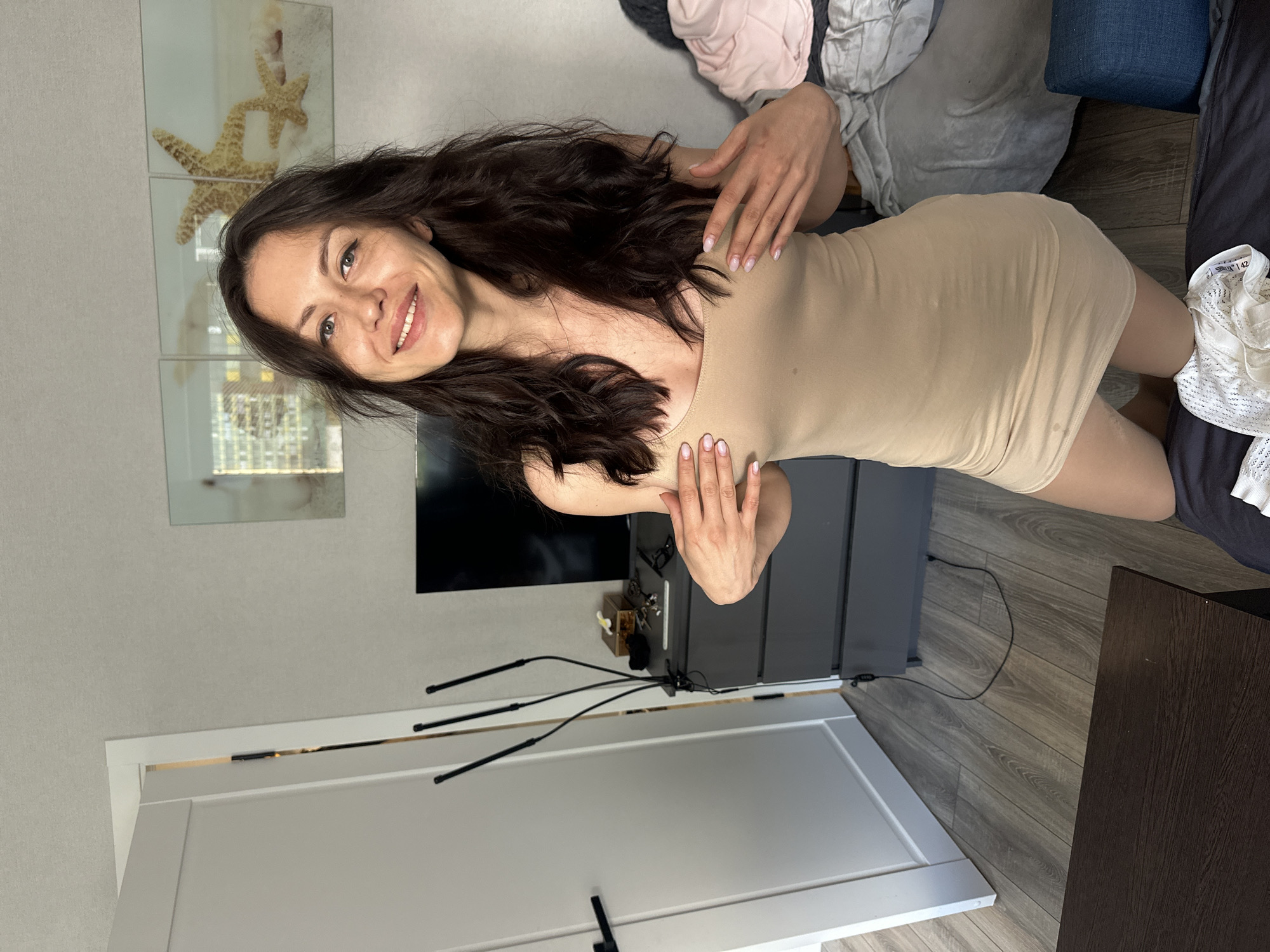 The image shows a woman with long, dark, wavy hair and a smiling expression. She is wearing a form-fitting, light beige dress. She is posing with her hands on her chest and hips, looking towards the camera.
The setting appears to be an indoor room. In the background, there is a dark dresser or cabinet, a television screen, and what looks like a wall with decorative artwork featuring starfish. There are also some clothes piled up in the background. To the left, there is a white door with black hardware. The flooring is a grey wood laminate.
The lighting suggests it is daytime, but no specific time of day can be determined. The overall mood seems relaxed and posed. There is no visible text in the image.