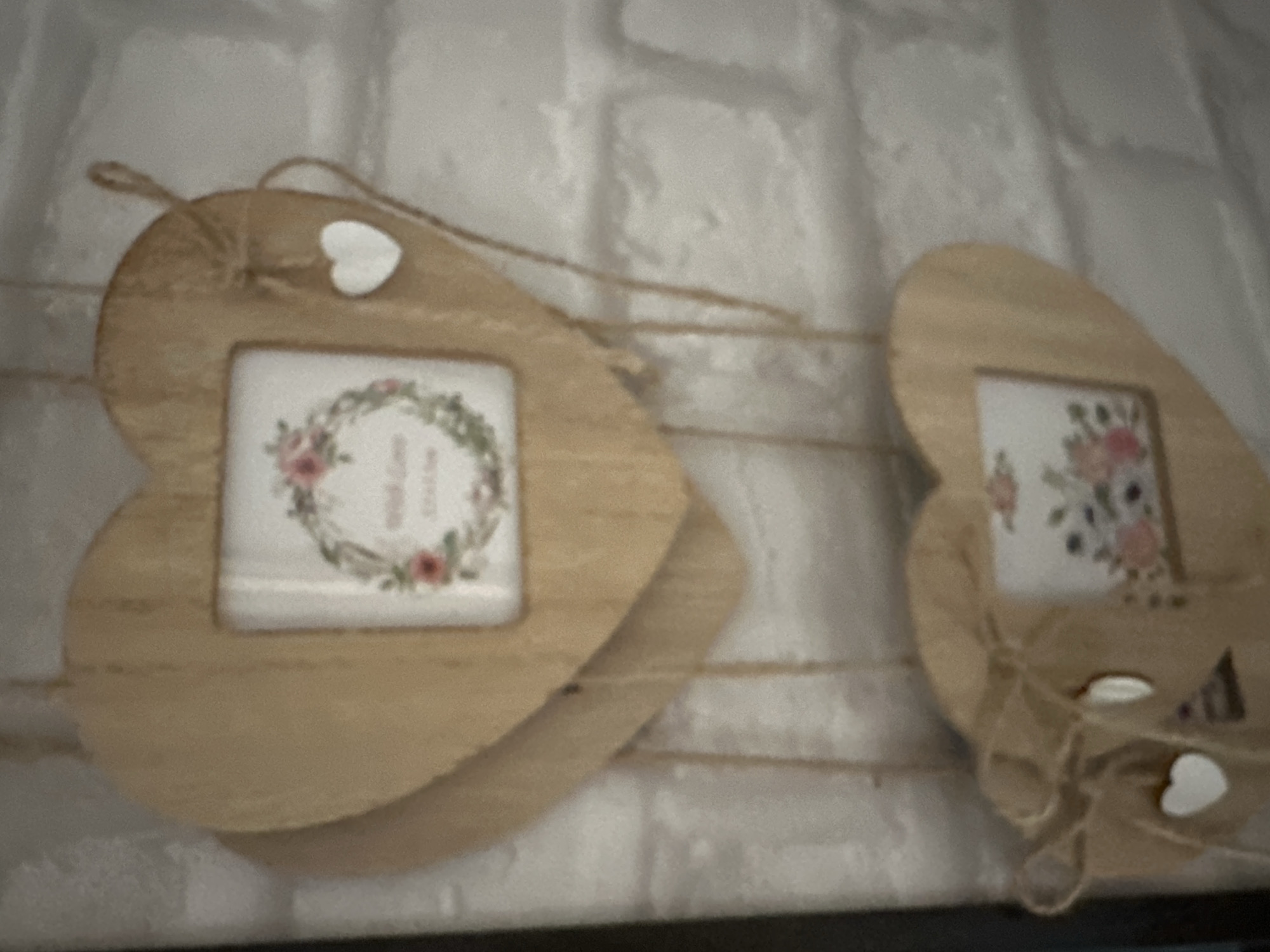 The image shows two heart-shaped wooden ornaments hanging against a white brick wall. Each ornament has a small square insert containing a floral print. The top ornament is larger and has a smaller white heart attached to its right side with twine. The bottom ornament is slightly smaller and has a bow made of twine tied to its left side, also with a small white heart hanging from it. The ornaments appear to be decorative items, possibly for a holiday or special occasion. There is no discernible location in Podolsk, Russia.