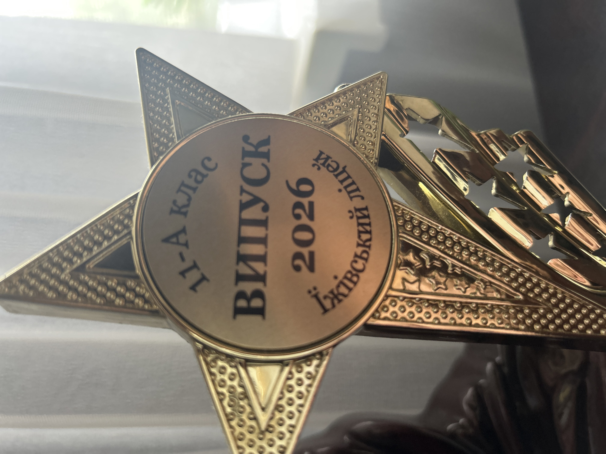 The image is a close-up shot of a gold-colored star-shaped trophy, likely awarded for academic achievement. The trophy features a circular medallion in its center, inscribed with Ukrainian text. The top arc of the medallion reads "11-А клас", indicating it's for the 11th grade, class A. Below this, in larger letters, is "ВИПУСК", meaning "Graduation". Underneath the graduation text is the year "2026". The bottom arc of the medallion bears the inscription "Їжівський ліцей", identifying the institution as the Yizhivtsi Lyceum. The trophy is highly detailed with embossed patterns and star cutouts on its pointed extensions. The background is blurred, showing white curtains and a hint of green foliage, suggesting an indoor setting near a window. The lighting is soft, casting a warm glow on the metallic surface of the trophy. There are no people or animals visible in the frame. The trophy is positioned on a dark, reflective surface, possibly a table or shelf, on which faint reflections of an unknown object can be seen.